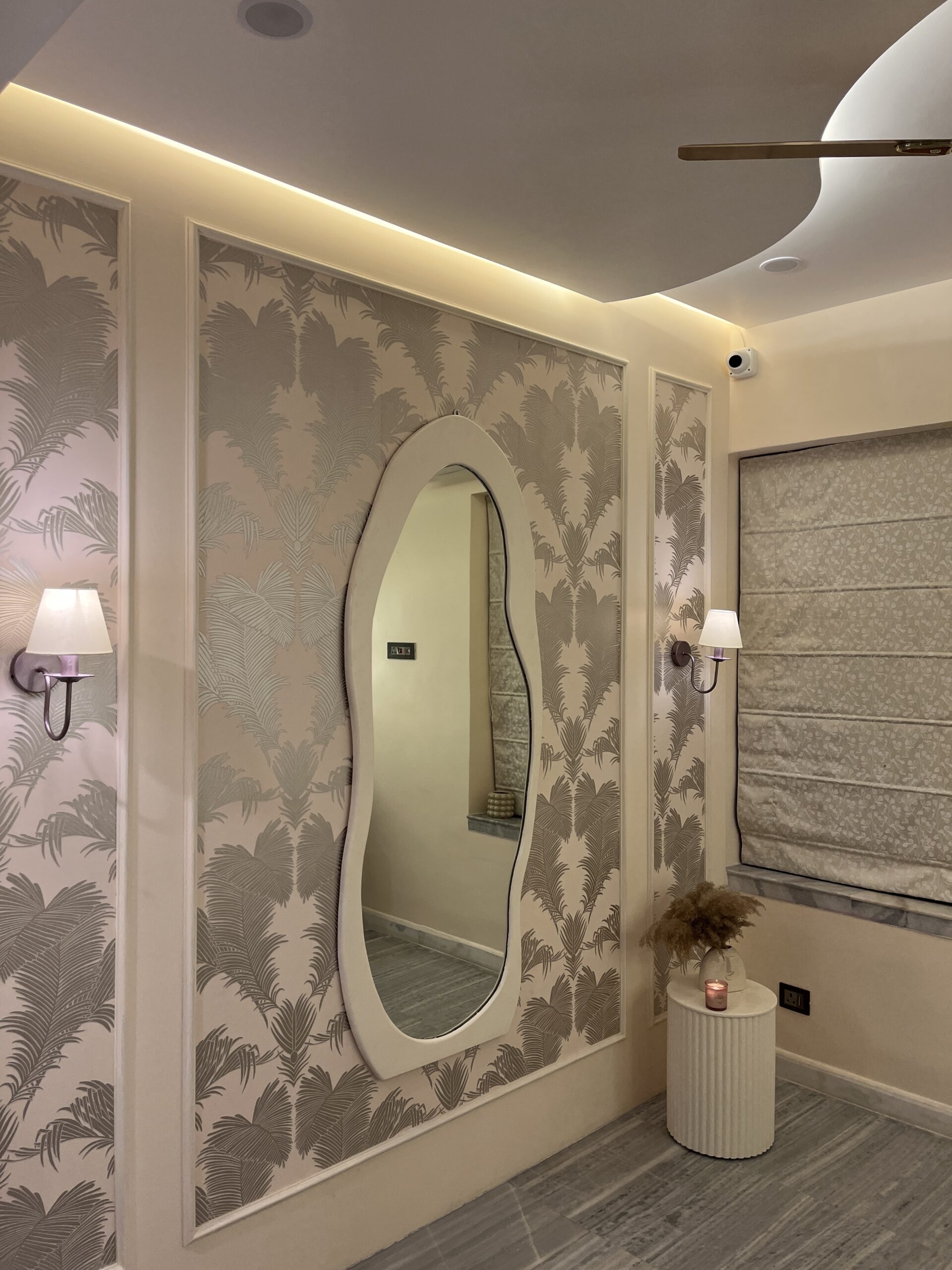 Modern bedroom accent wall featuring tropical palm leaf wallpaper framed by classic white wall mouldings, a large wavy asymmetrical full-length mirror, elegant wall sconces, and a fluted side table