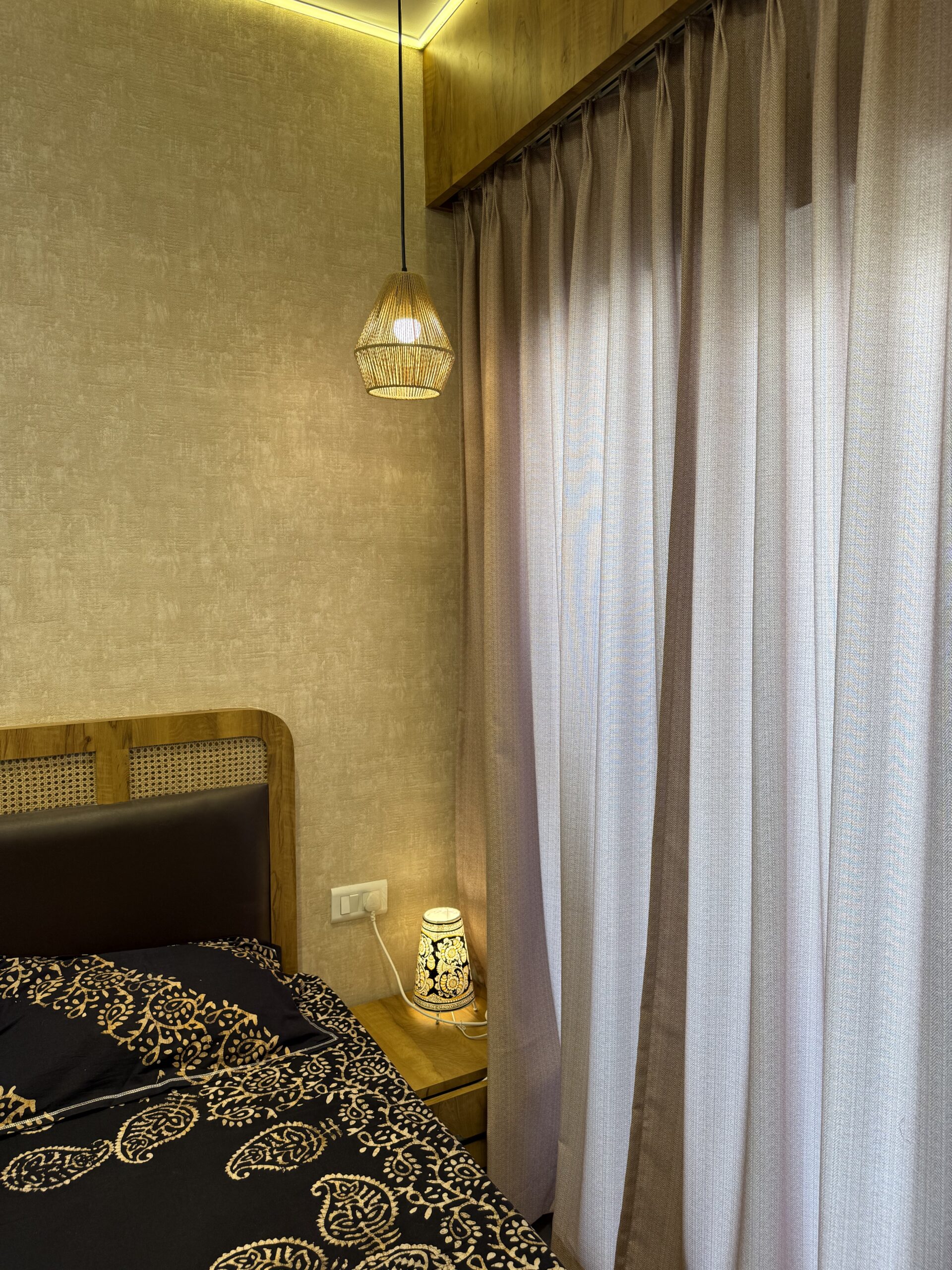 Bedroom bedside corner featuring a wooden headboard with cane detailing, textured beige wallpaper, layered lighting with a wicker pendant lamp, and neutral floor-to-ceiling sheer curtains.