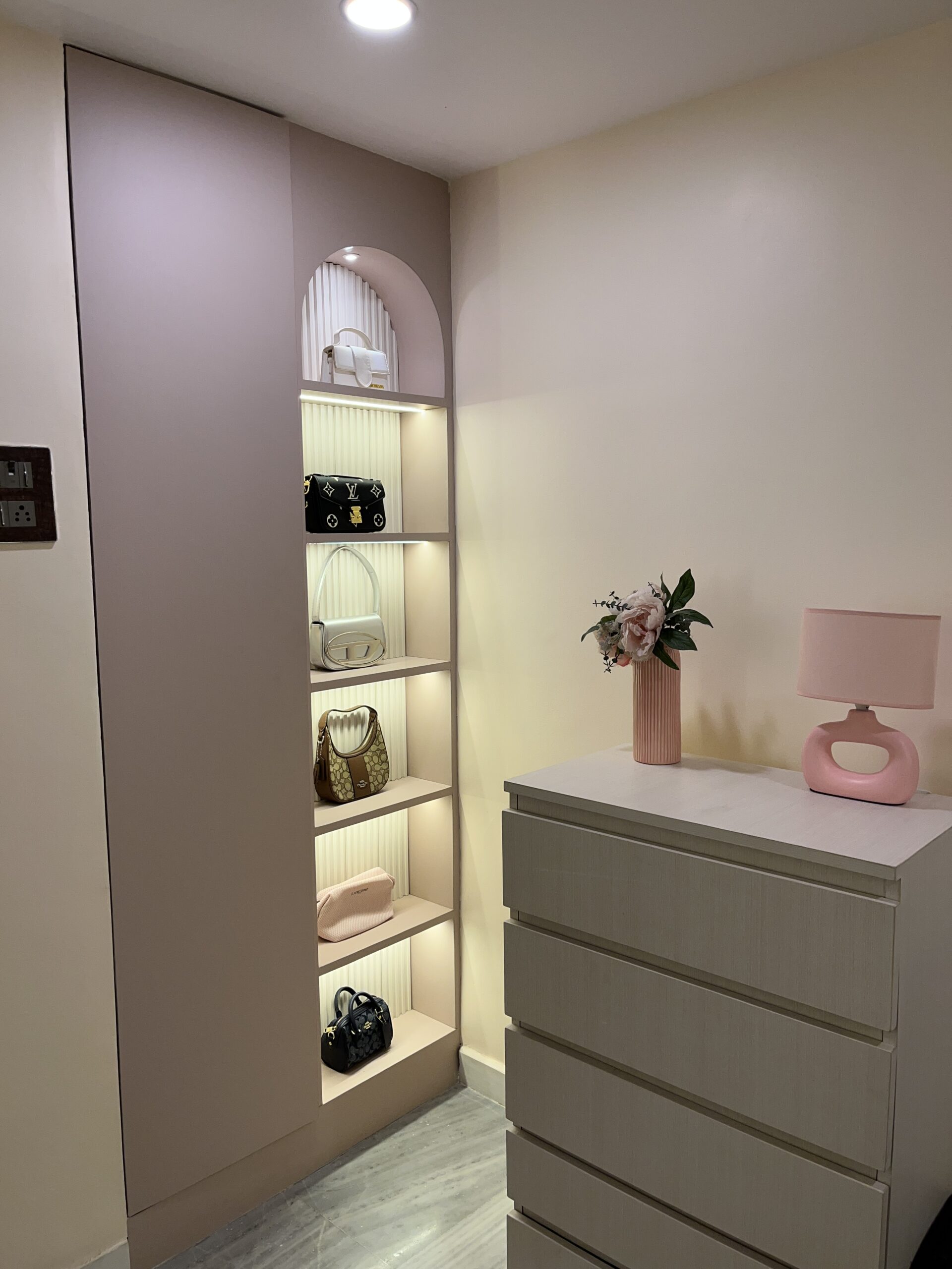 Modern walk-in closet featuring a dusty rose wardrobe wardrobe unit with an integrated arched open display shelf, vertical fluted back paneling, warm internal LED strip lighting showcasing luxury handbags, and an adjacent cream-colored chest of drawers