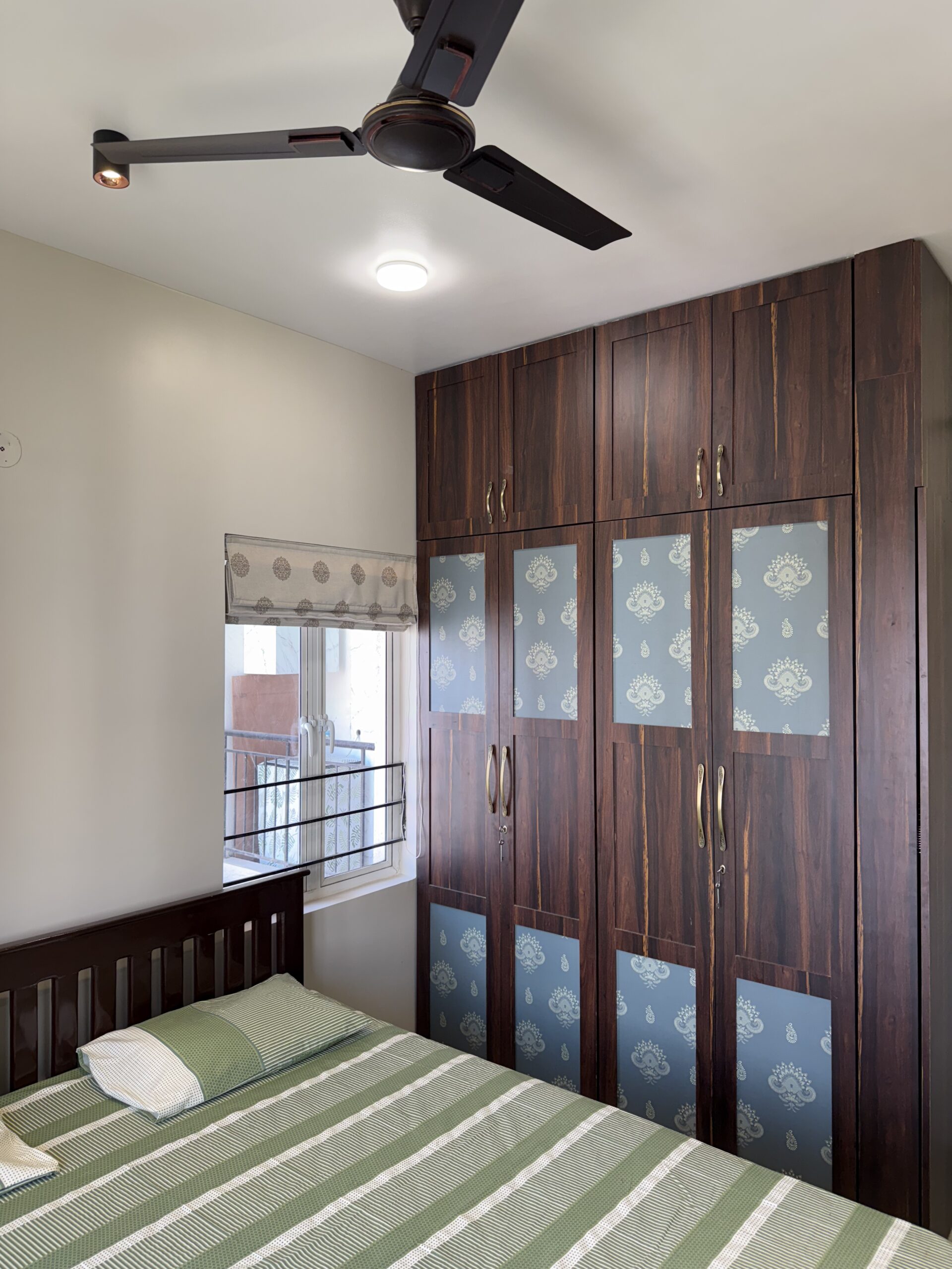 Modern floor-to-ceiling bedroom wardrobe made of dark wood laminate featuring laminates with intricate traditional patterns, antique brass handles, and an overhead loft storage system.
