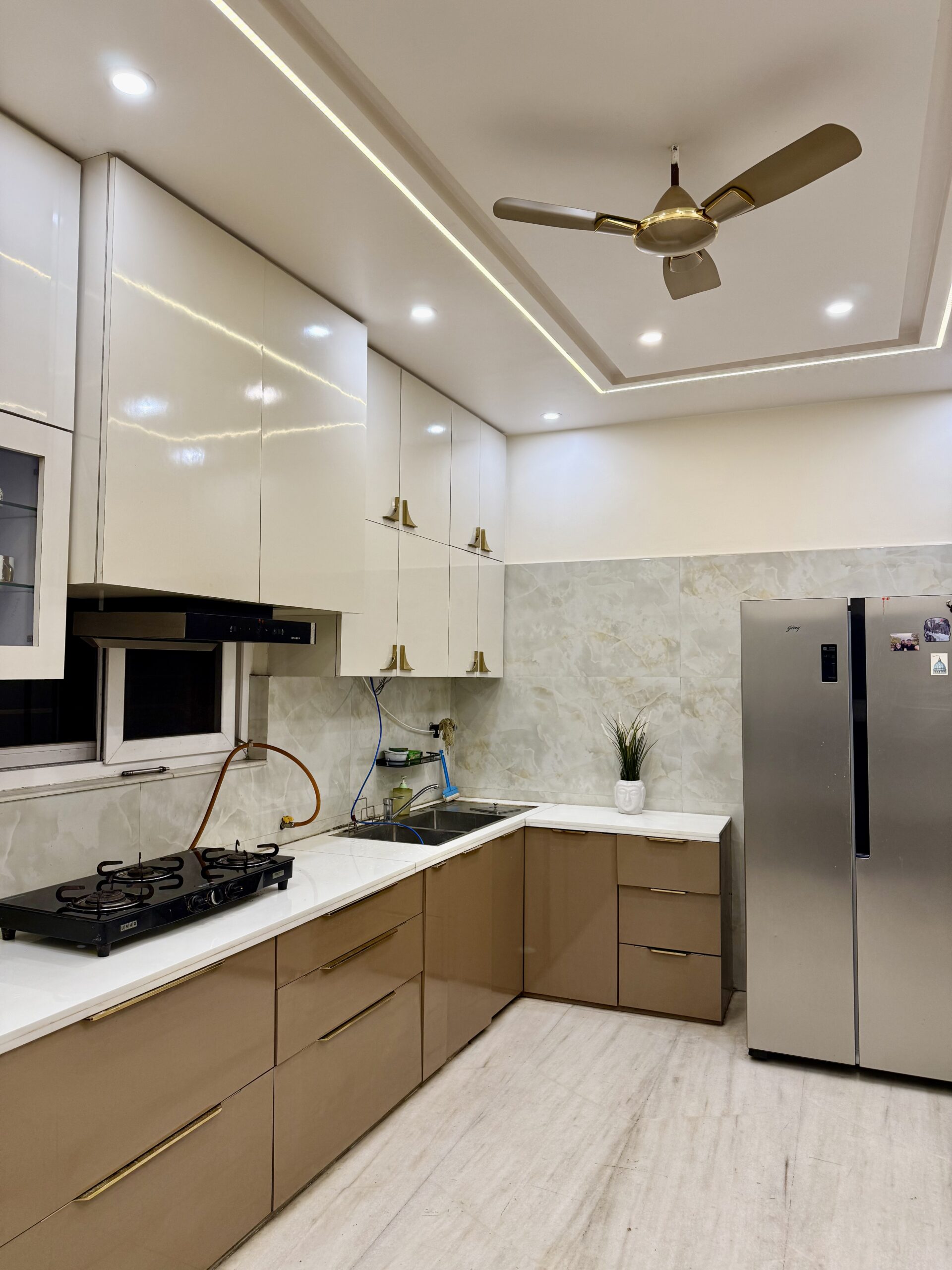 Modern L-shaped modular kitchen featuring high-gloss white upper cabinets with gold handles, warm beige lower drawers, white countertops, an integrated gas stovetop, a double-bowl sink, marble-patterned wall tiles, and a false ceiling with linear LED profile lighting