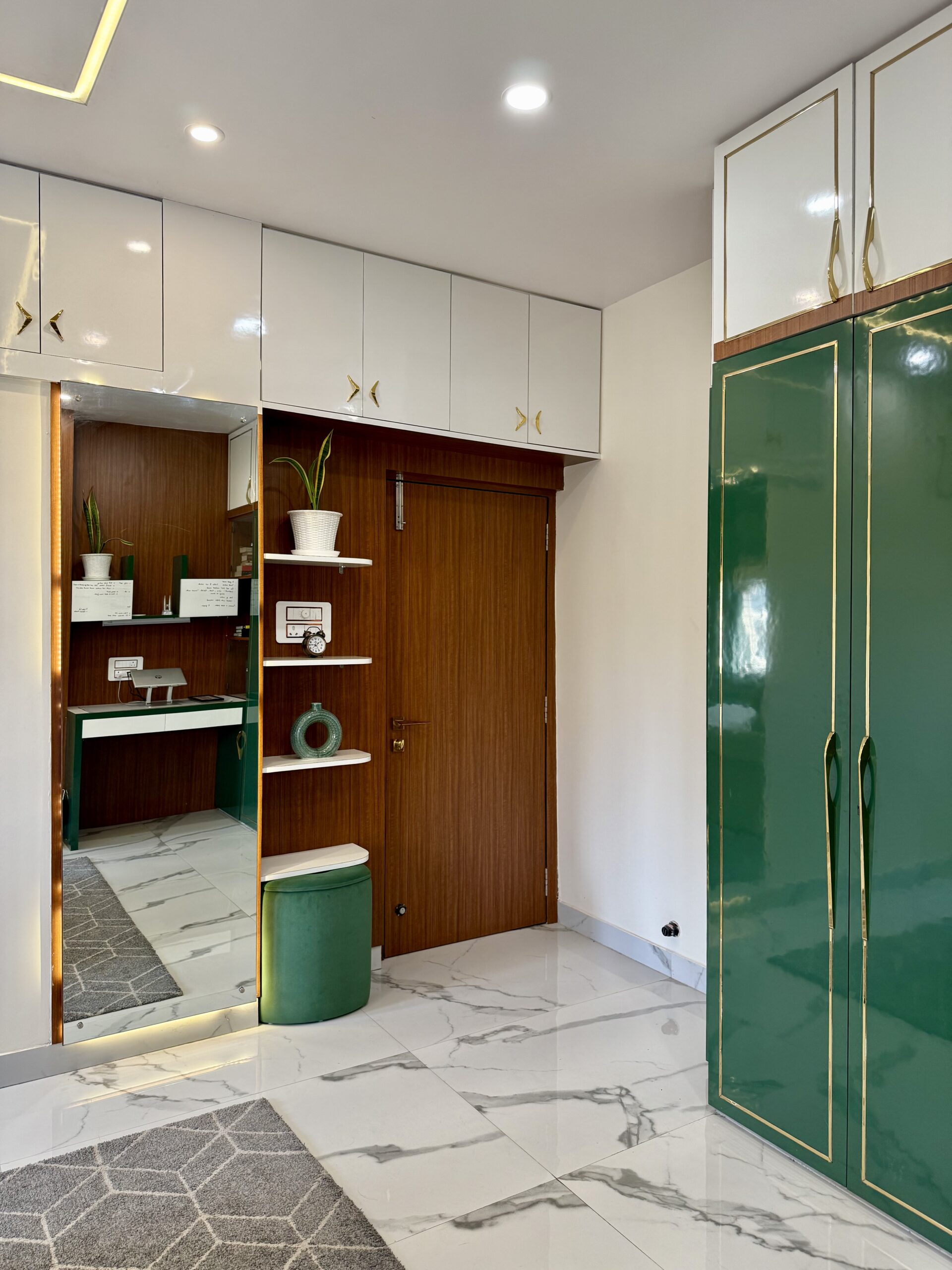Modern bedroom interior showcasing high-gloss emerald green wardrobes with gold inlay handles, white upper loft cabinets, a seamless wood-paneled entryway door with display shelves, a full-height dressing mirror, and polished marble flooring
