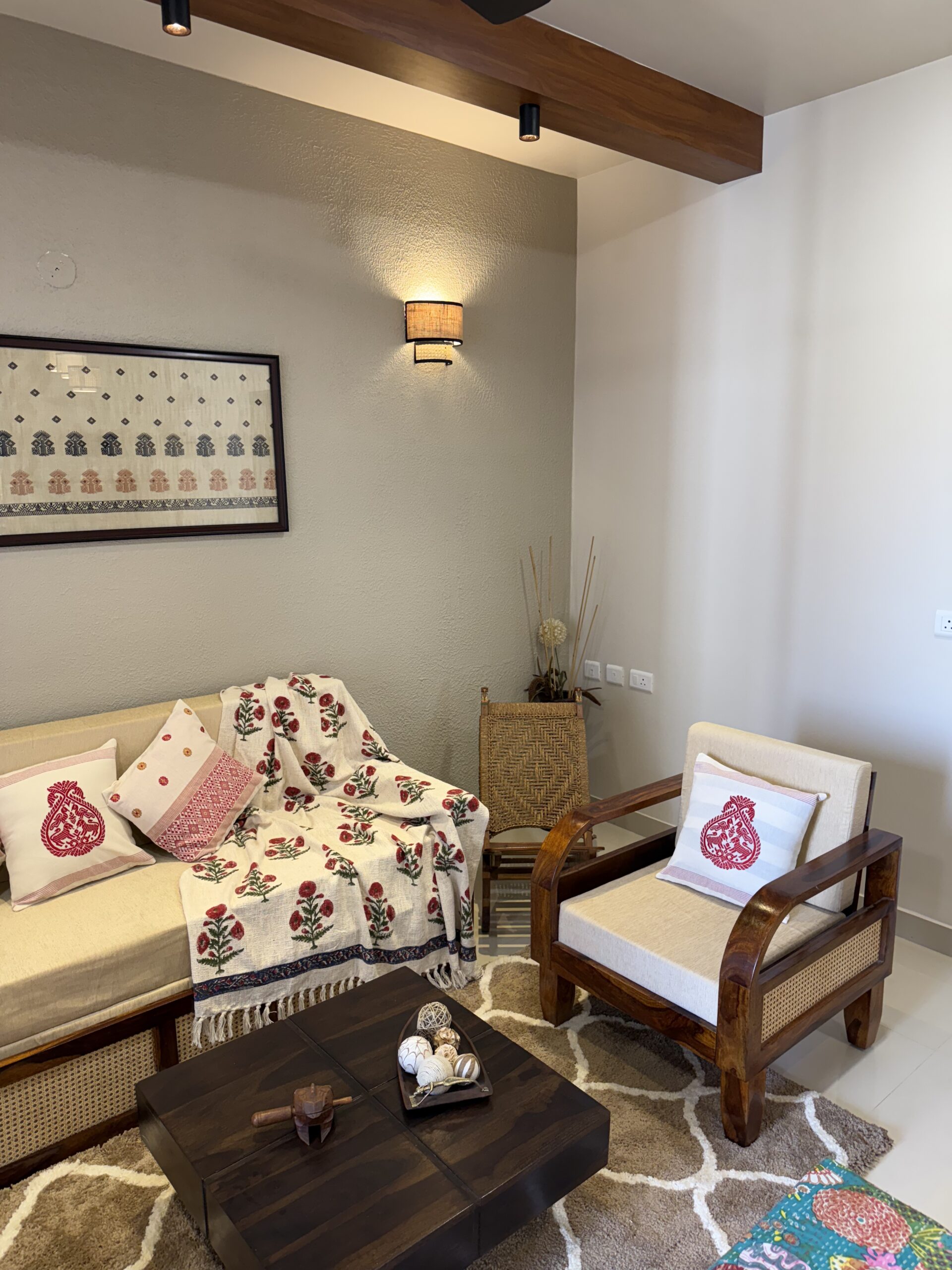 Modern Indian living room featuring a wooden sofa and matching accent armchair with woven cane details, traditional block-printed textiles, a dark wooden coffee table, textured grey wall, and a rustic woven sconce light.