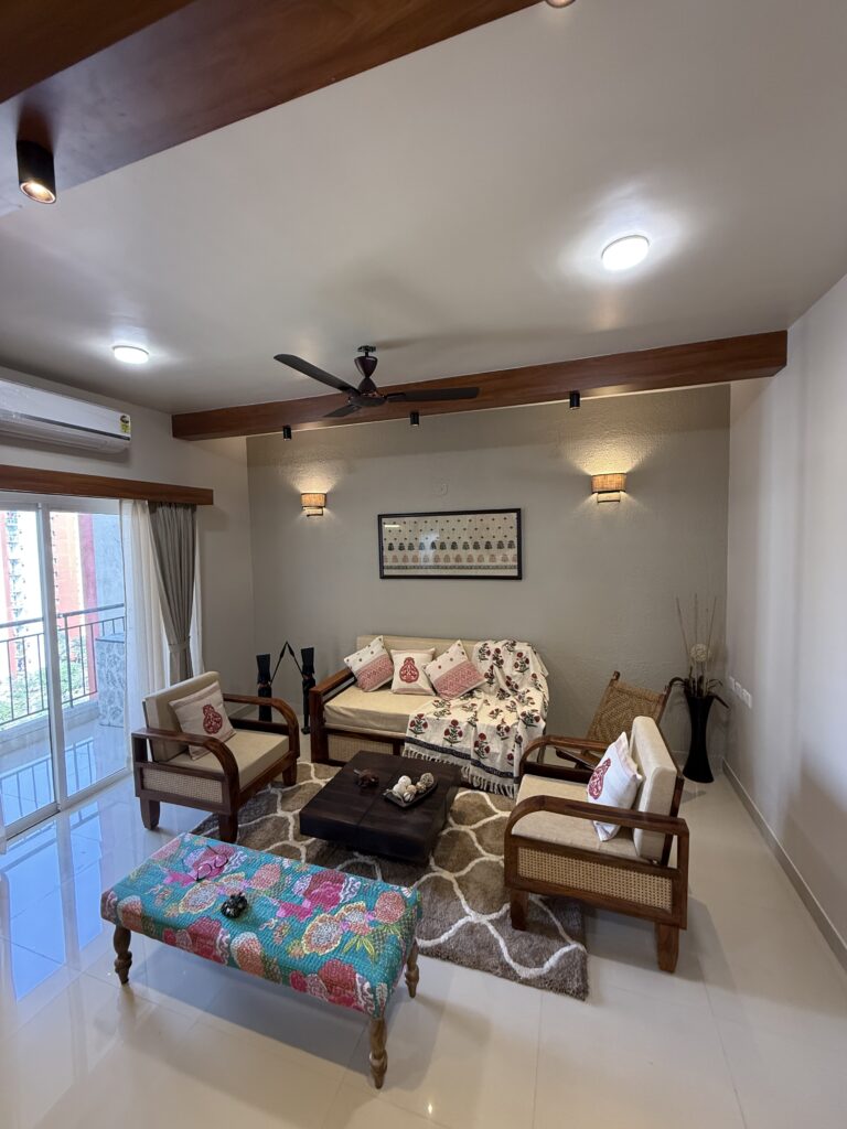 Full view of a modern Indian living room featuring wooden and cane furniture, an ethnic patterned bench, a plush geometric area rug, textured accent wall with traditional framed art, and a false ceiling with prominent wooden beams and spotlights.