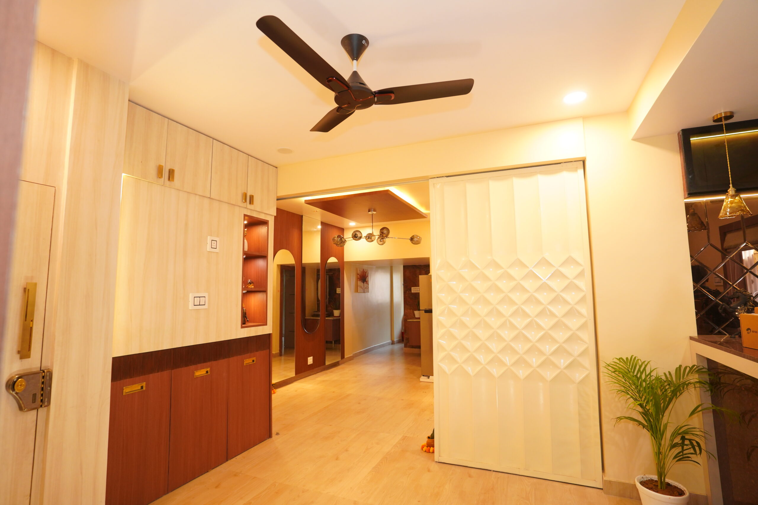Modern apartment interior view showing an entrance foyer with a light wood storage cabinet, an ivory sliding partition door with a 3D geometric diamond pattern, a false ceiling with a ceiling fan, and a long hallway leading to a dining area with a modern chandelier