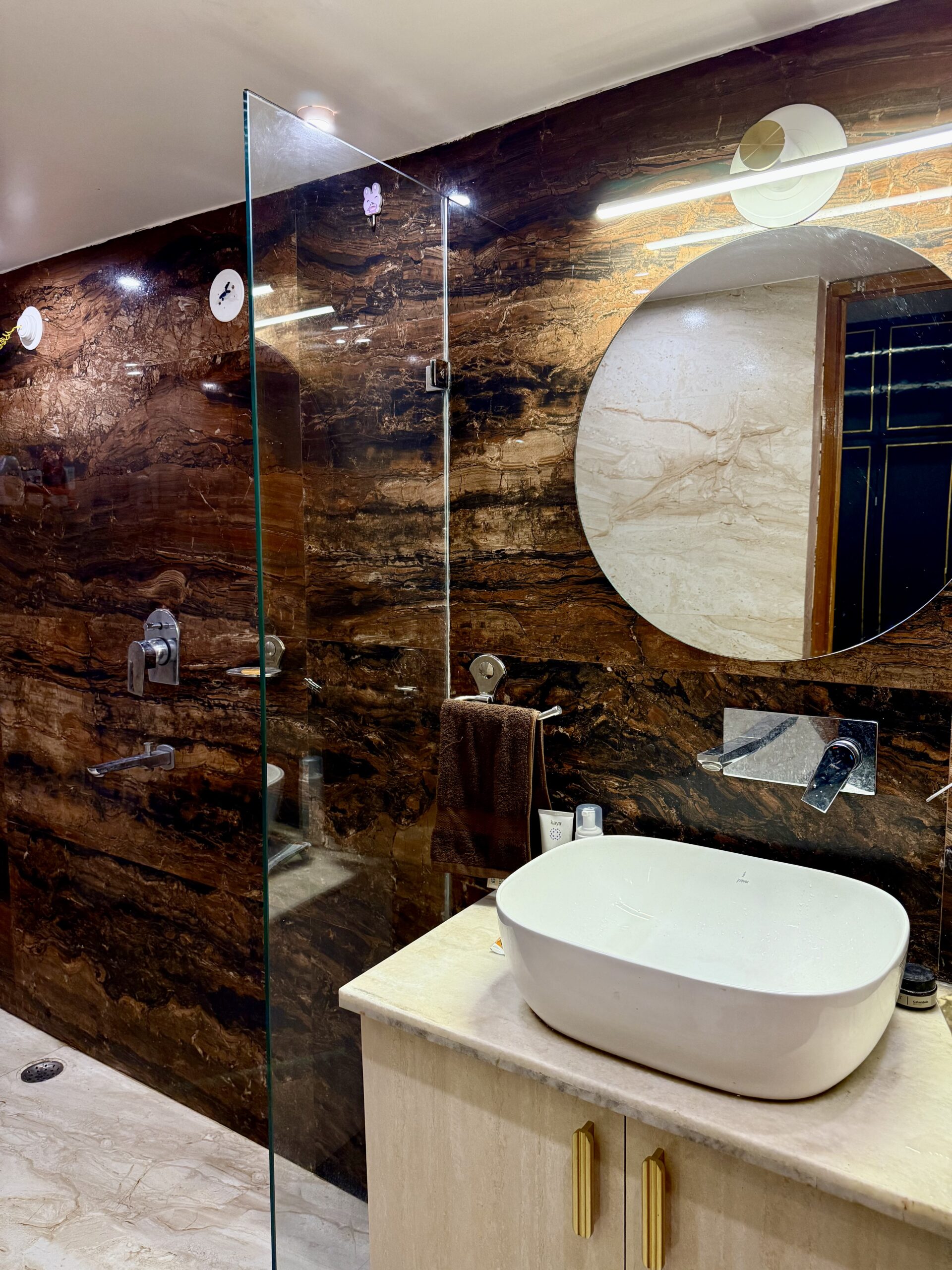 Luxury bathroom interior featuring dramatic dark brown marble-textured wall tiles, a modern glass walk-in shower partition, a round wall mirror with an overhead linear light, and a white countertop basin on a light wood vanity with gold handles