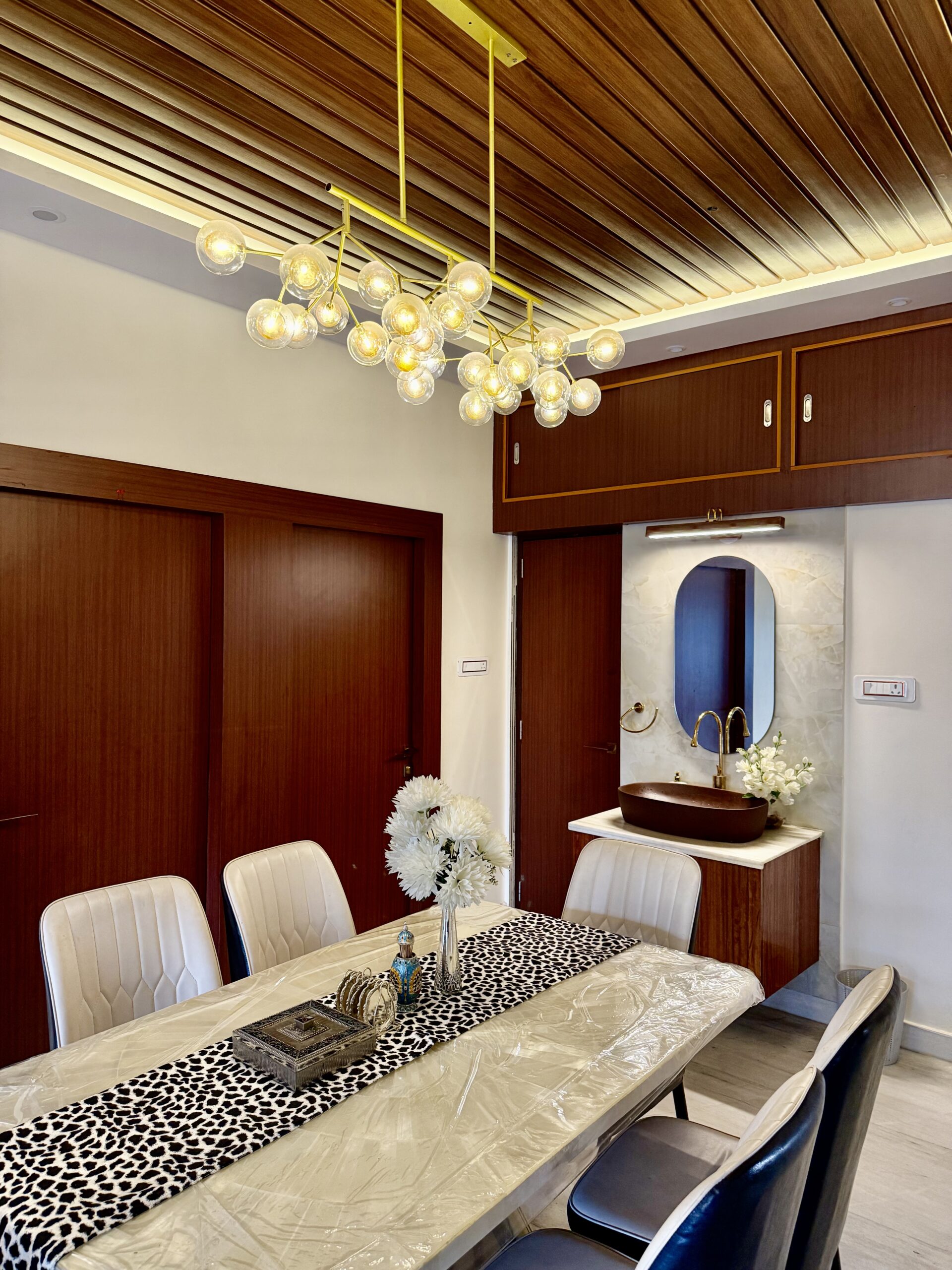 Luxury dining room featuring a modern multi-globe glass chandelier, a wooden fluted panel false ceiling, a white marble dining table with tufted leather chairs, and an elegant floating washbasin vanity with a capsule mirror against a marble-veined accent wall