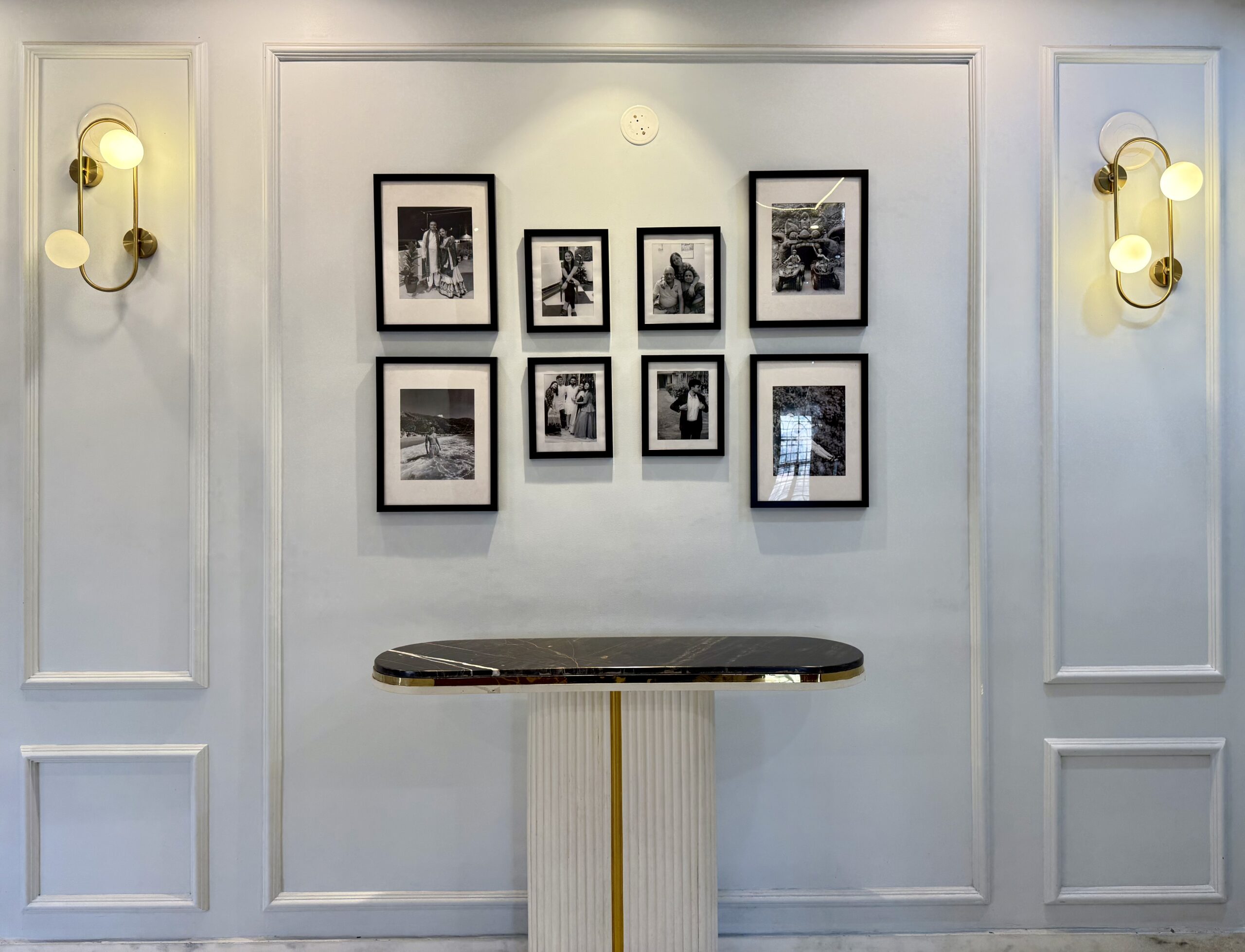 Symmetric luxury foyer featuring a black-and-white grid photo gallery wall framed by classic white wainscoting trim, a fluted white pedestal console table with a black marble top, and two modern brass loop globe sconces