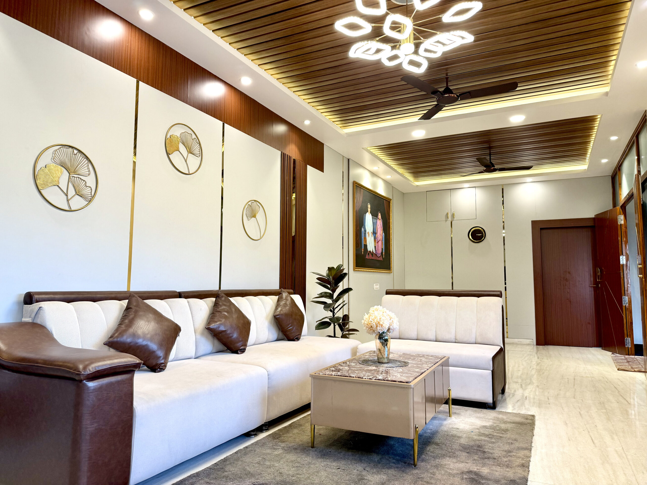 Grand open-concept living hall featuring white accent wall panels with gold metal trim, circular brass ginkgo leaf wall art, dual-tone leather sofas, a marble-top coffee table, and an intricate wooden rafter false ceiling with a modern geometric chandelier