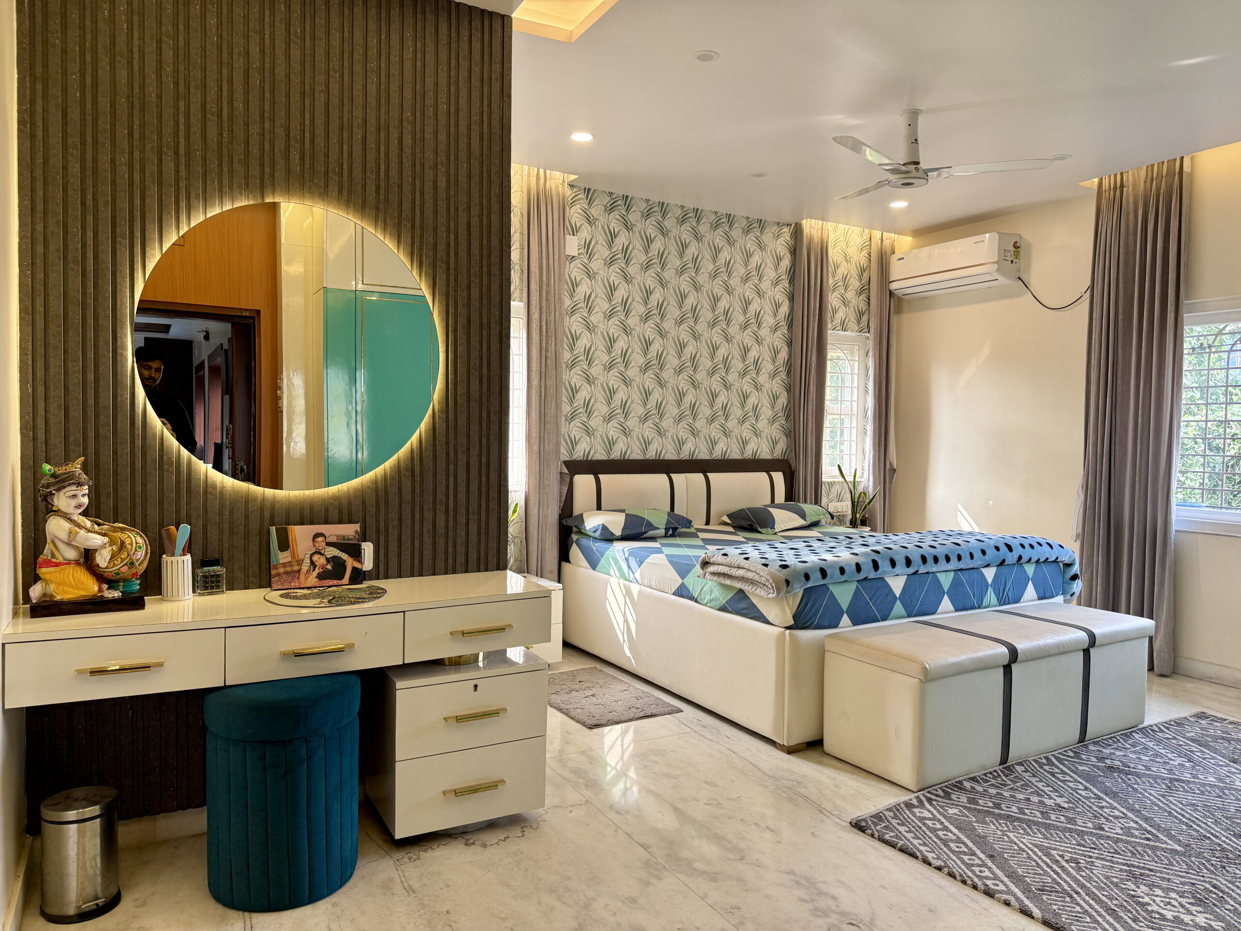 Spacious master bedroom featuring a white vanity table with gold handles, a large round backlit LED mirror on a dark grey fluted panel wall, a teal velvet stool, a double bed with a matching storage bench, and a tropical leaf-patterned accent wallpaper wall