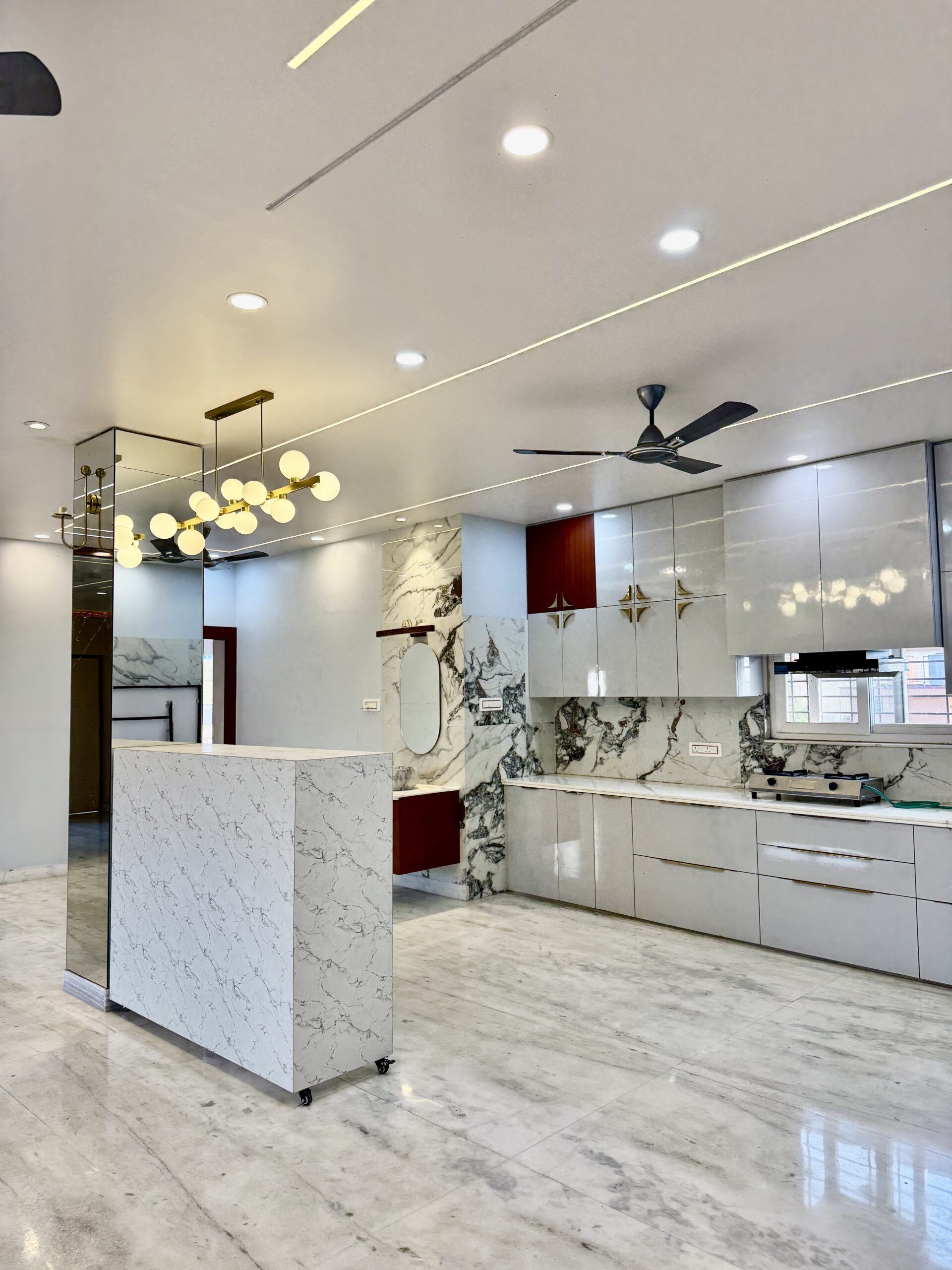 Luxury open-plan modular kitchen with high-gloss white cabinets, marble-patterned backsplash and flooring, a matching marble-textured breakfast island counter, an elegant gold sputnik globe chandelier, and seamless false ceiling profile lighting