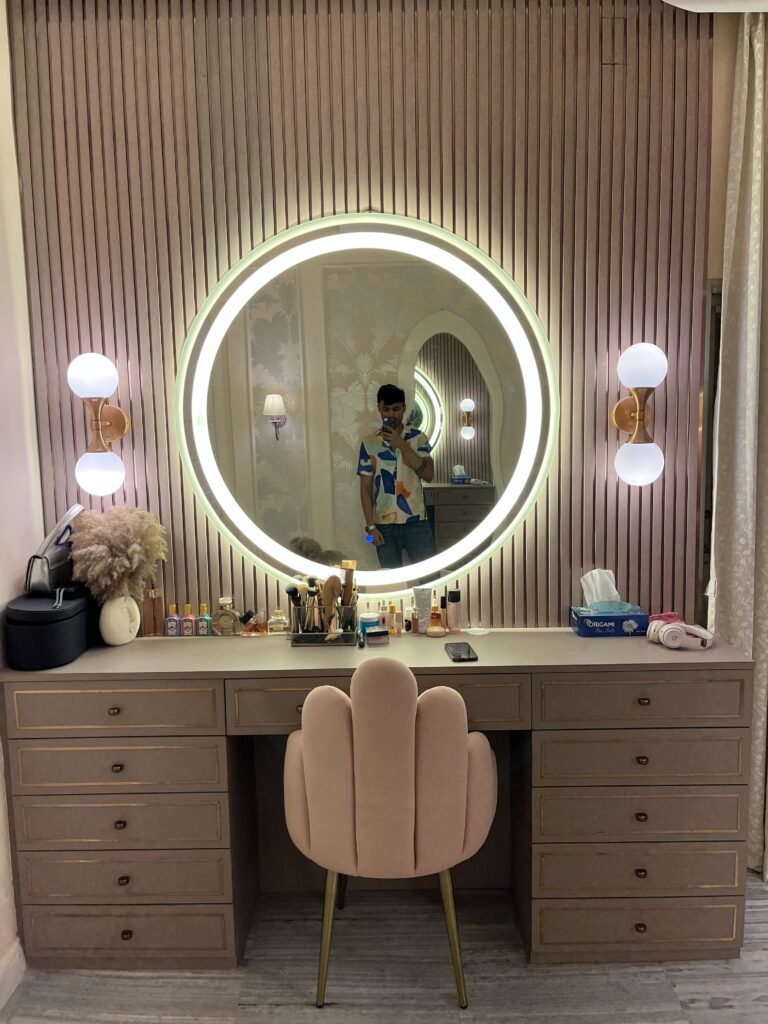 Luxury vanity dressing table with gold inlay drawers, a large round backlit LED smart mirror, dusty pink fluted wall paneling, modern brass globe sconces, and a plush scalloped pink velvet chair