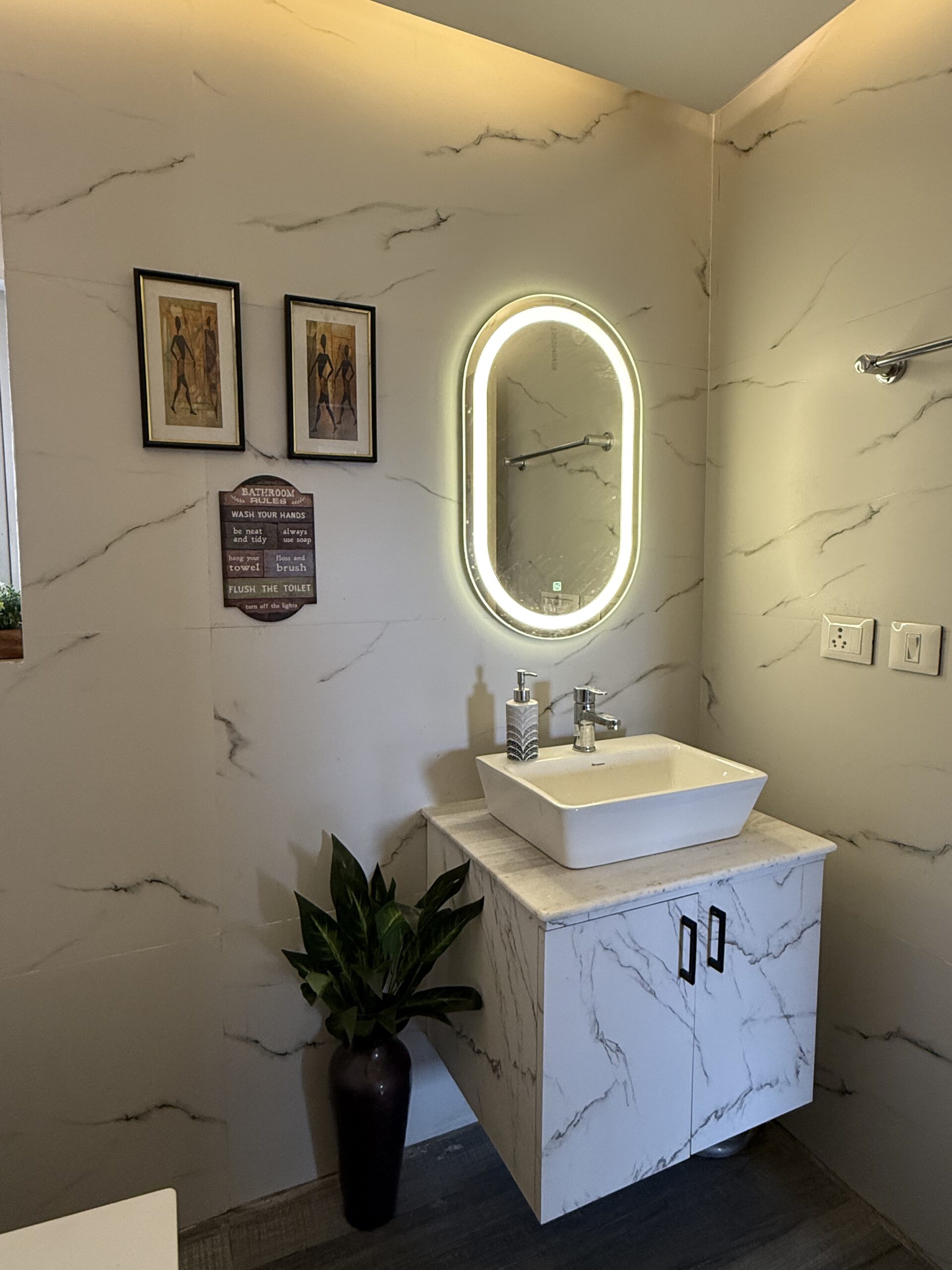 Modern bathroom interior featuring a floating marble-look vanity cabinet, a white tabletop washbasin counter, an illuminated capsule-shaped LED back-lit mirror, and matching white marble-textured wall tiles.