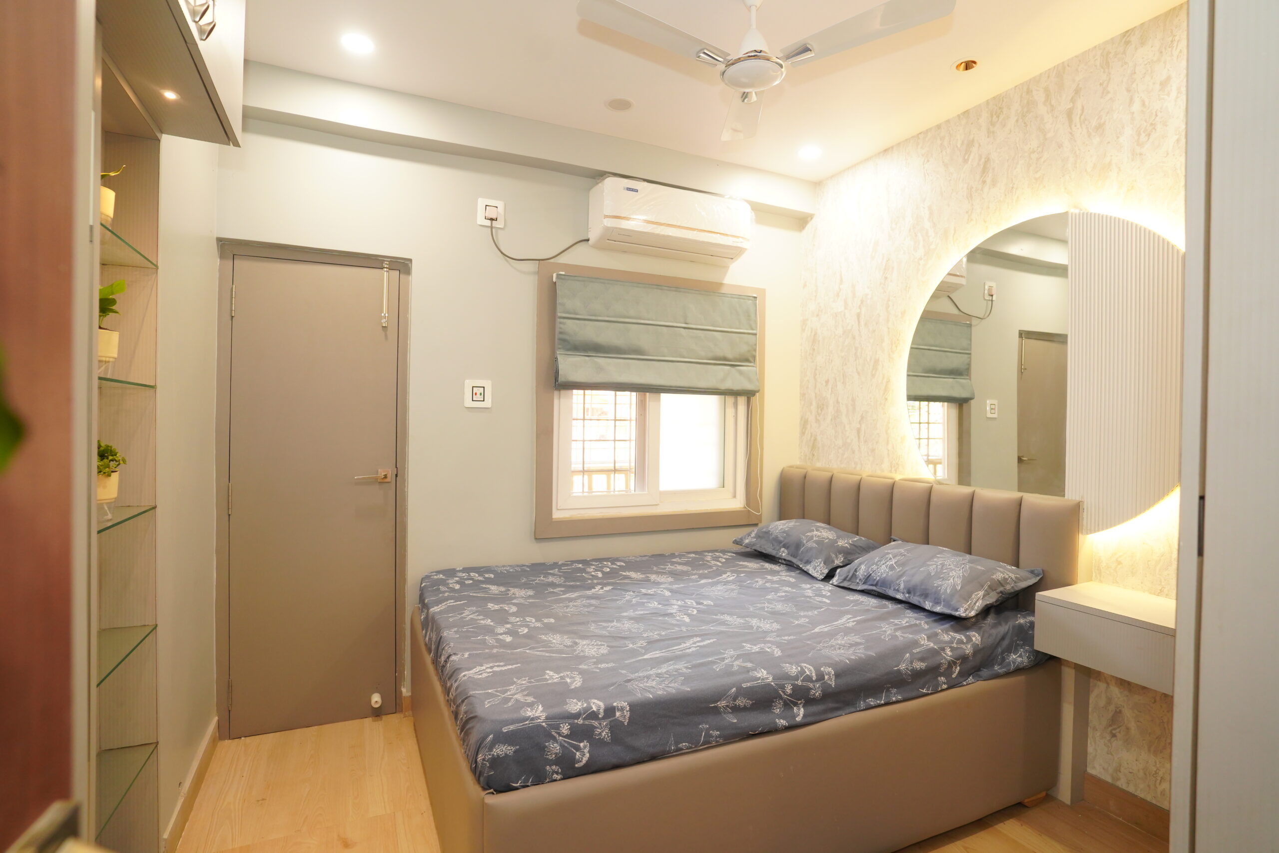Modern bedroom interior featuring a double bed with a grey channel-tufted headboard, a textured accent wallpaper wall, a large backlit semi-circular mirror flanked by white fluted paneling, a sage green Roman window blind, and glass display shelving