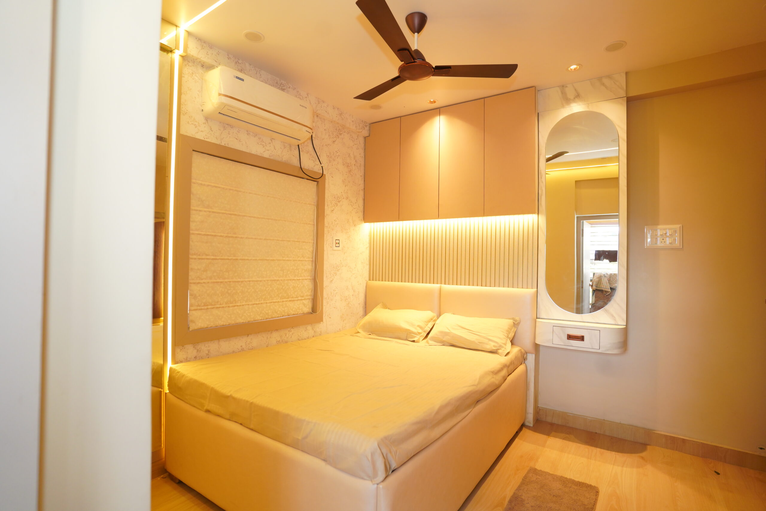 Modern bedroom interior featuring a double bed with a plush upholstered headboard, an overhead storage loft cabinet with custom fluted paneling below it, built-in warm LED accent strip lighting, a Roman window blind, and a floating capsule-shaped vanity mirror