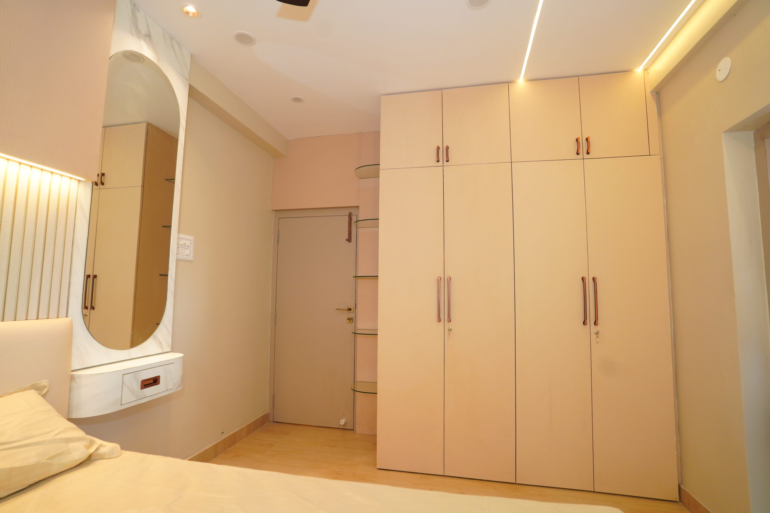 Modern bedroom interior featuring a large beige floor-to-ceiling modular wardrobe with top lofts, a sleek capsule-shaped wall-mounted vanity mirror with marble-textured framing, and a false ceiling with a recessed linear LED profile light