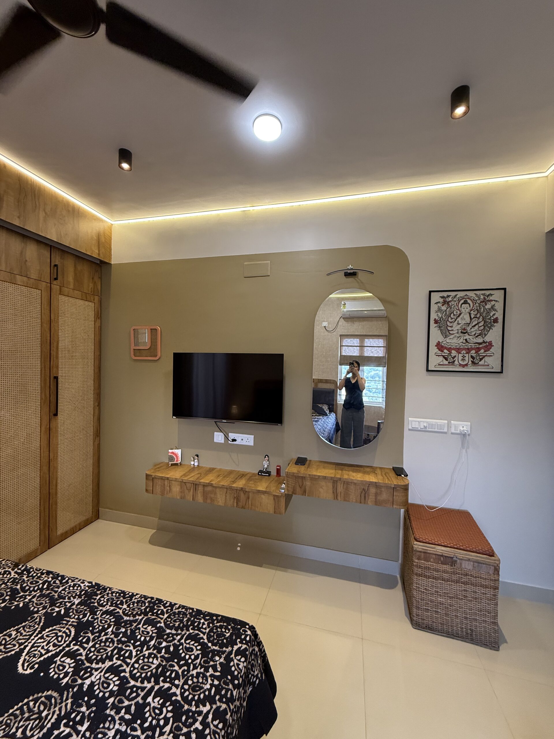 Modern bedroom interior featuring a floating wooden TV console, a matching vanity unit with an capsule oval mirror, a wardrobe with cane panels, and warm false ceiling LED strip lighting.