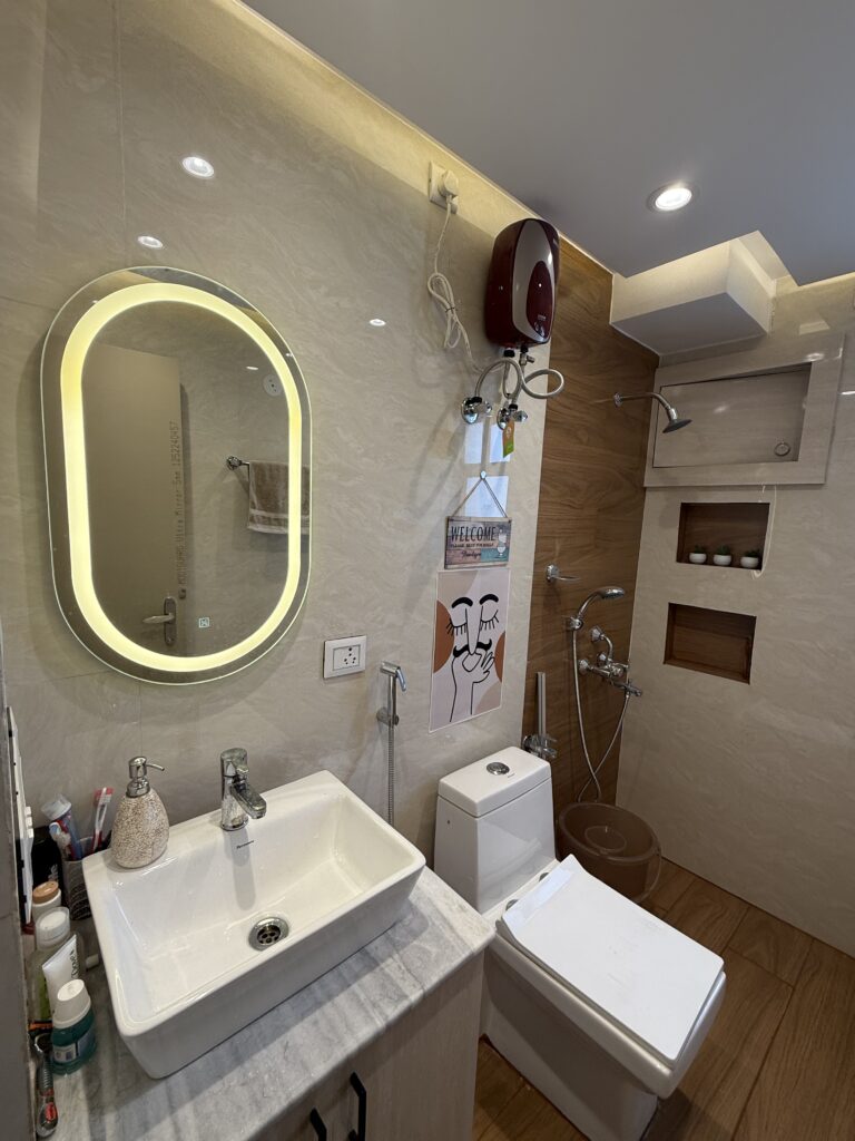 Compact bathroom interior with glossy beige wall tiles, a wooden accent wall behind the shower area, an oval LED backlit mirror over a countertop washbasin, a modern commode, and built-in wall niches.