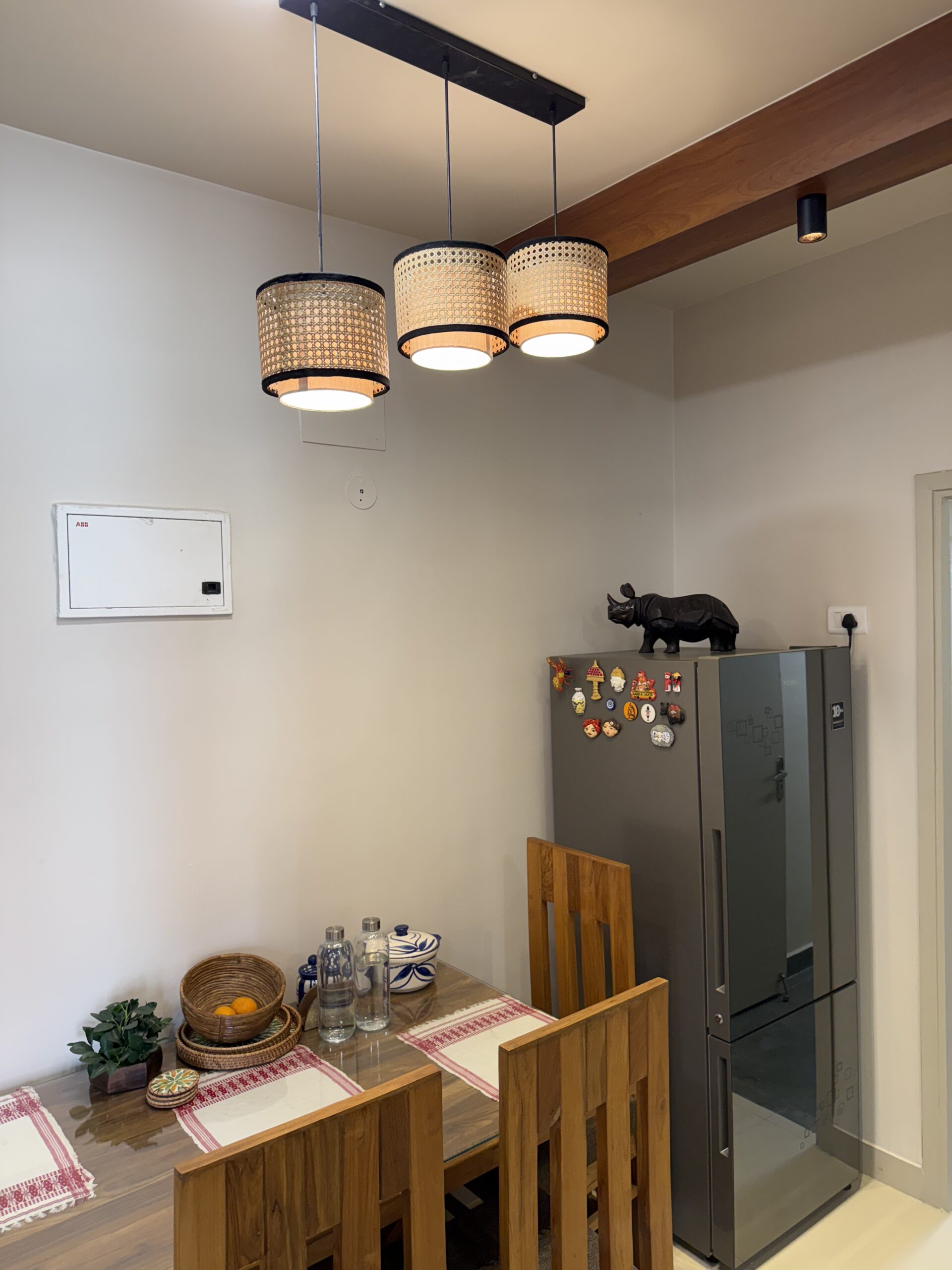Modern dining room area featuring a wooden dining table and chairs, a triple-cluster woven cane pendant light fixture, a false ceiling with a wooden beam, and a double-door refrigerator.