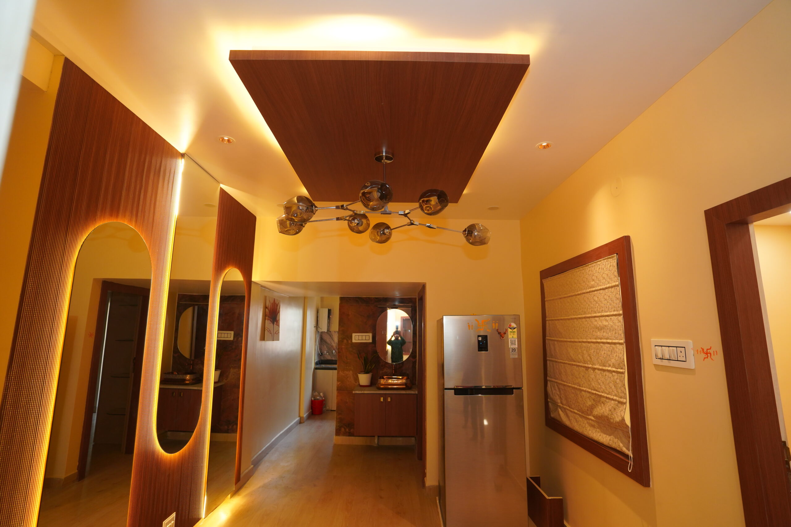 Modern dining space featuring a wooden island false ceiling panel with warm cove lighting, a contemporary branch-style glass globe chandelier, a full-height mirror wall with back-lit arched cutouts, and a double-door refrigerator