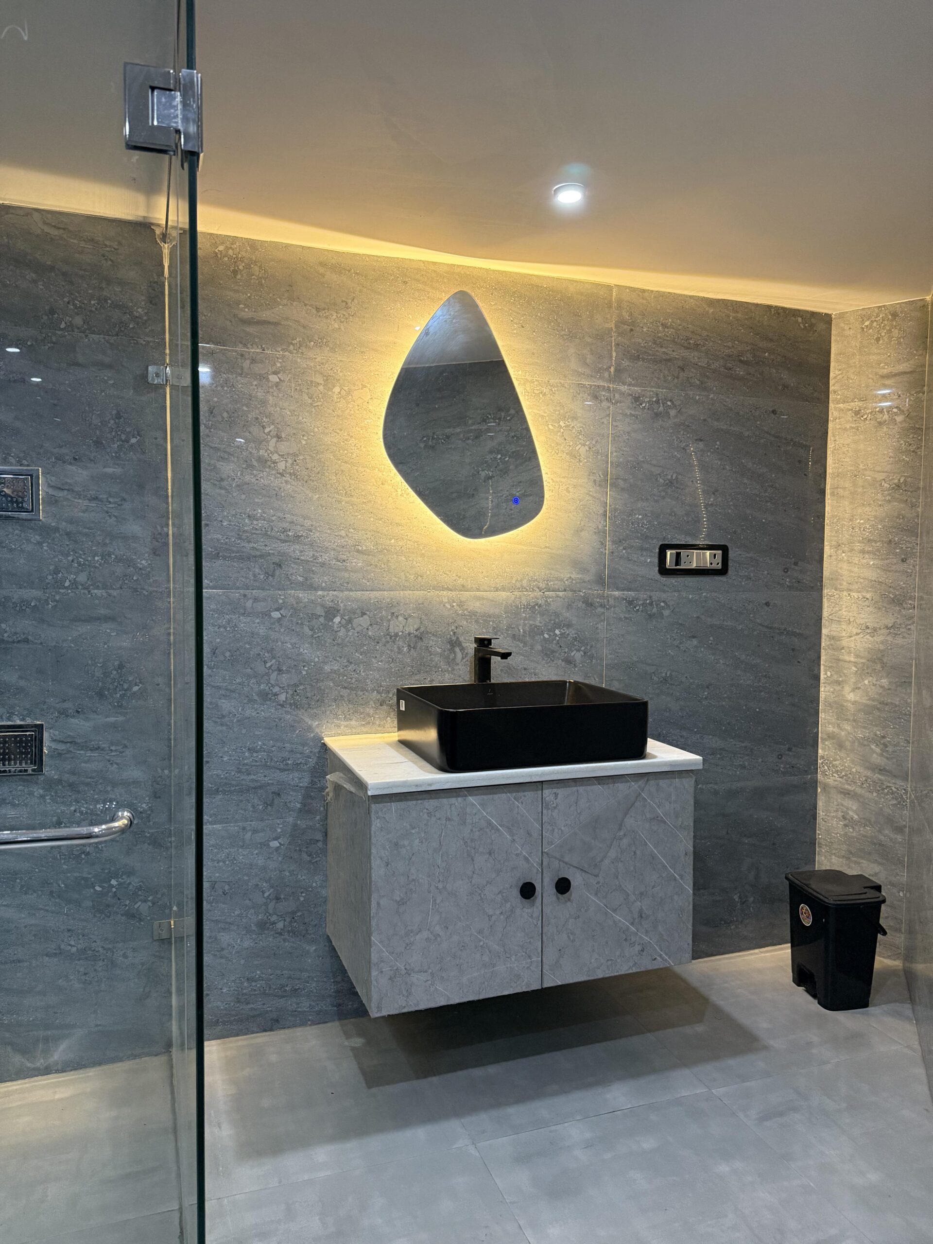 Modern bathroom interior featuring a floating grey stone-textured vanity cabinet, a matte black countertop washbasin with a black faucet, an organic asymmetrical backlit LED mirror with warm illumination, and large-format grey marble-patterned wall tiles