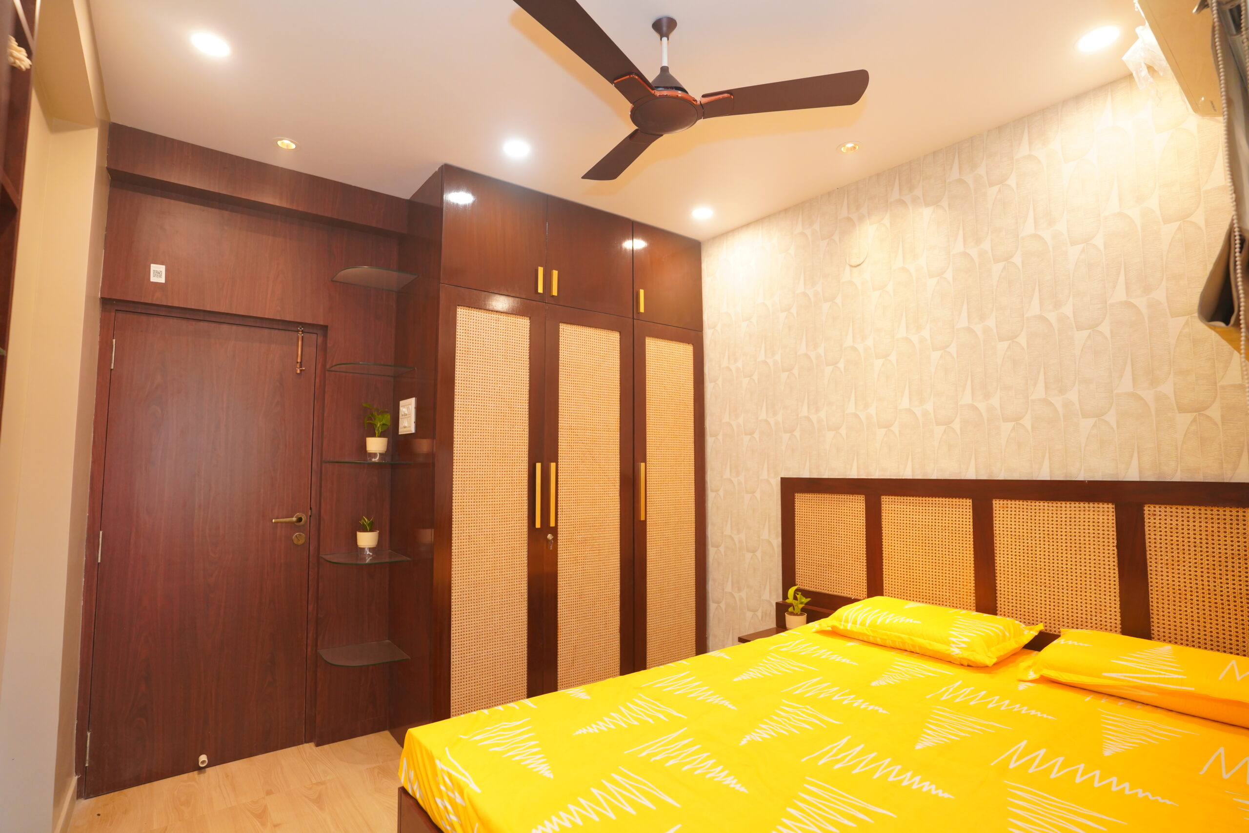 Modern guest bedroom featuring a dark wood wardrobe with woven cane door panel inserts, a matching dark wood headboard with cane paneling, subtle patterned wallpaper, glass corner display shelves, and bright yellow bedding