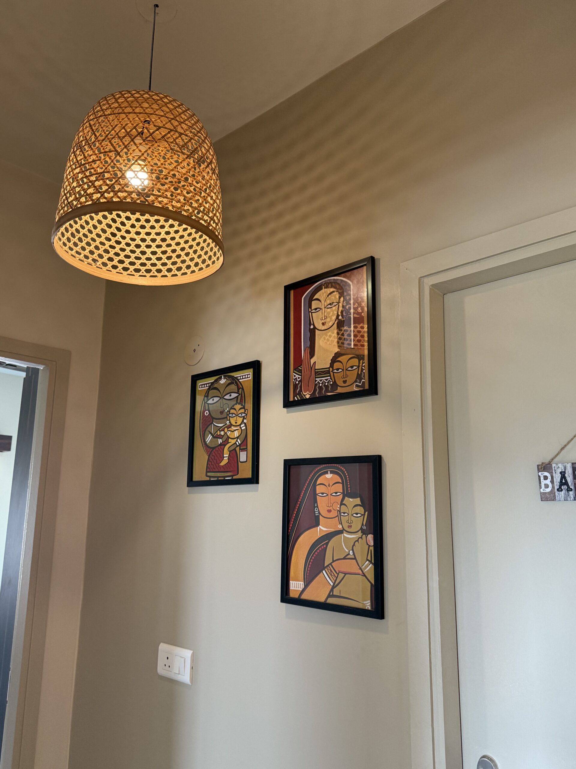 Home corridor featuring a gallery wall with three framed traditional Indian folk art paintings, a woven bamboo basket pendant light casting intricate mesh shadows on the ceiling, and warm neutral walls.