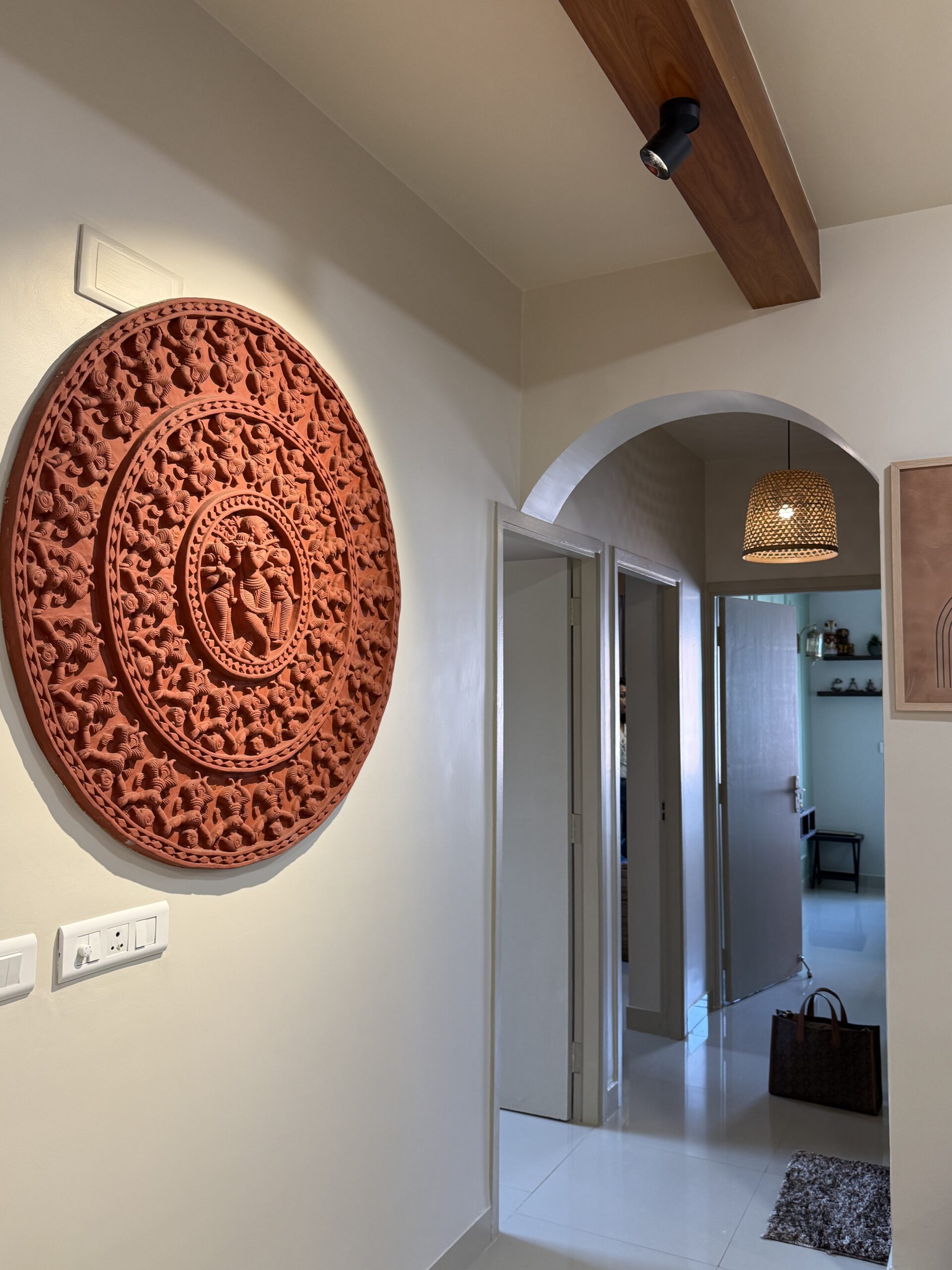 Modern home hallway and corridor featuring a large circular terracotta wall mural with intricate traditional Indian carvings, an arched doorway, and a wooden ceiling beam with a black spotlight.