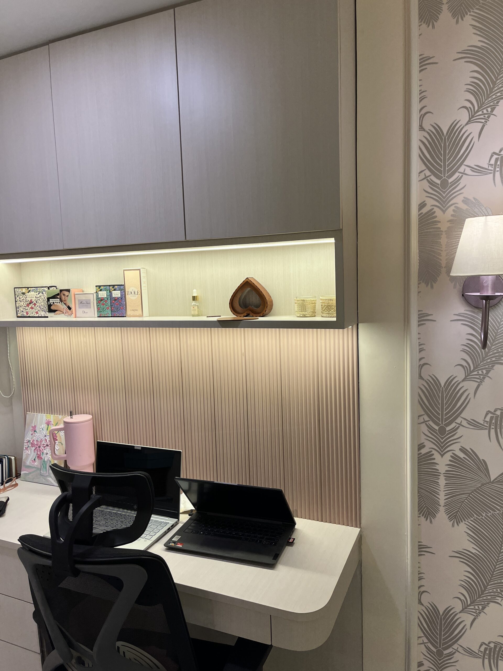 Modern home office workspace featuring a light-colored wooden desk, pink fluted wall paneling, overhead storage cabinets with an integrated LED backlit open display shelf, laptops, an ergonomic office chair, and an adjacent tropical wallpaper accent wall