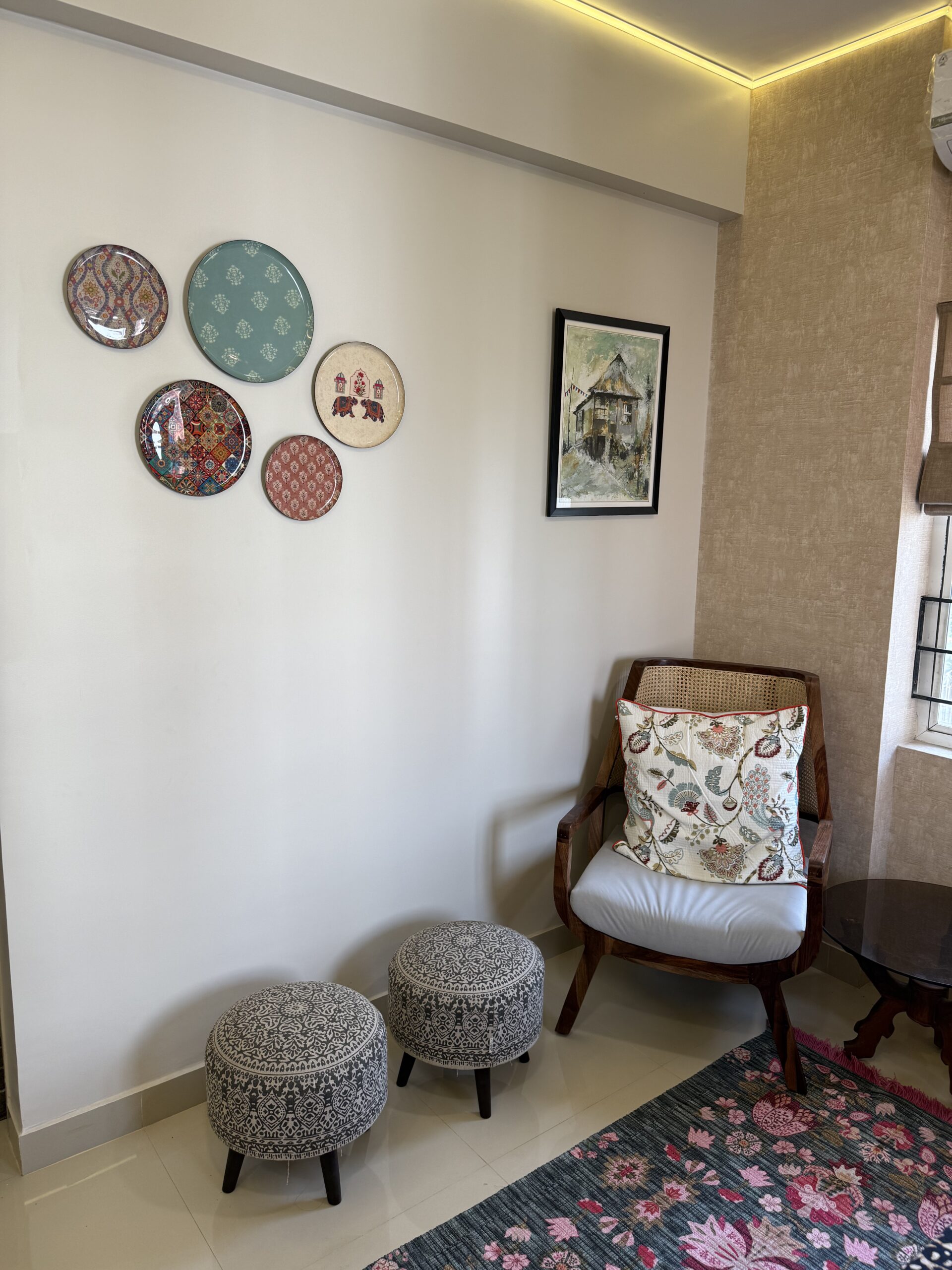Cosy sitting corner featuring a wooden accent armchair with cane backrest, patterned pouf ottomans, decorative ceramic wall plates, and warm false ceiling LED lighting.