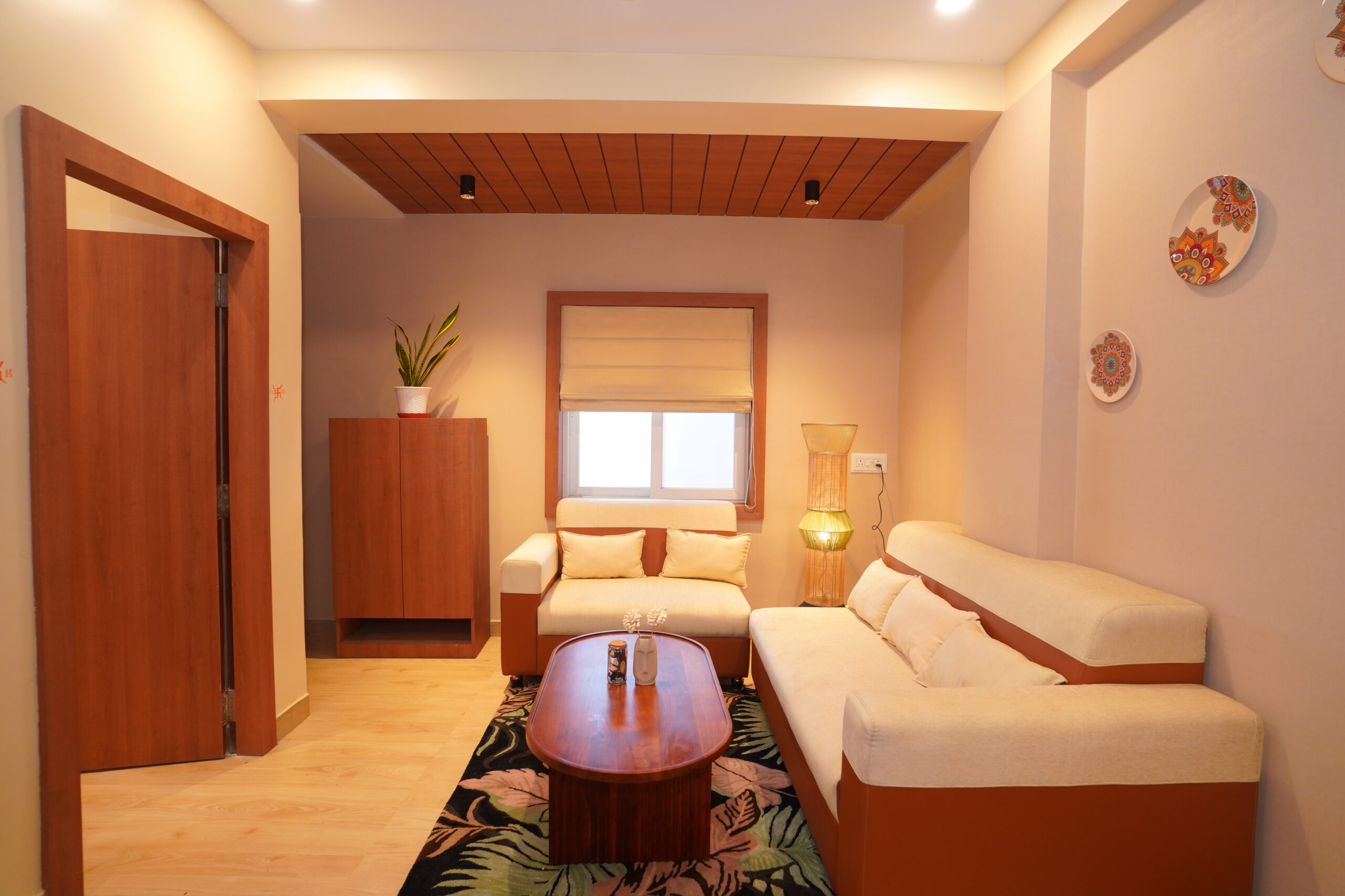 Modern living room featuring a dual-tone cream and tan sofa set, an oval wooden coffee table, a tropical patterned area rug, a distinct wooden false ceiling section with black spotlights, a Roman window blind, and decorative wall plates