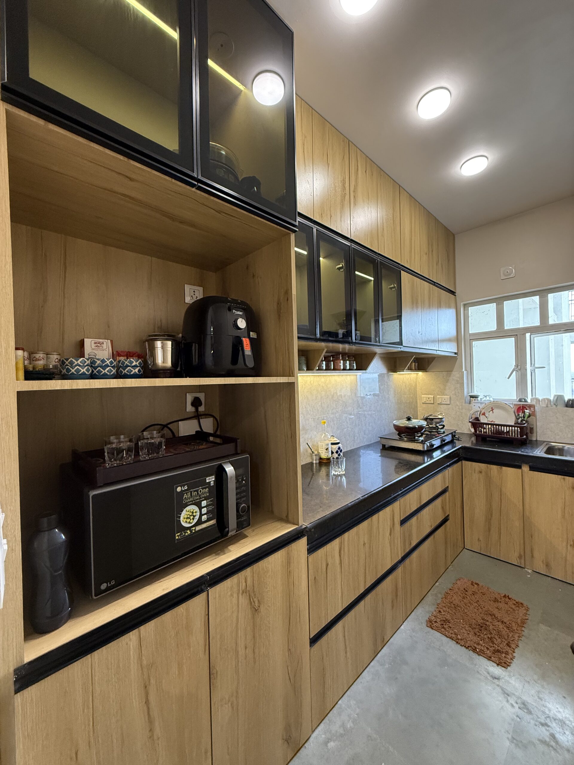 Modern L-shaped modular kitchen featuring light wood laminate cabinets, black profile handles, upper fluted glass cabinets with LED strip lighting, dark granite countertops, an integrated appliance console housing a microwave and air fryer, and a built-in stovetop.