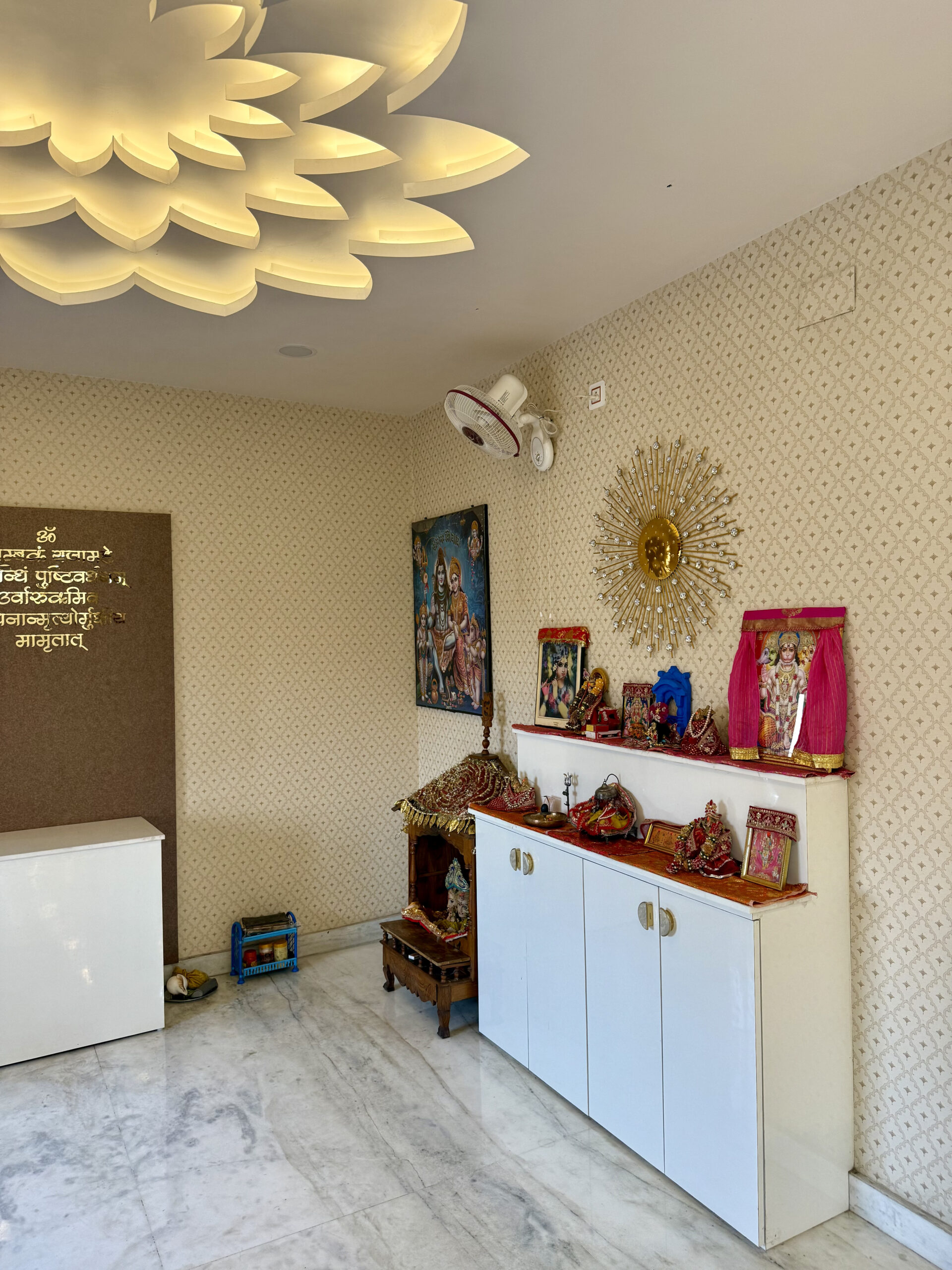 Pooja room interior featuring a spectacular multi-layered lotus flower false ceiling with warm hidden cove lighting, patterned wallpaper, marble flooring, custom storage cabinets, and brass spiritual script on a textured wall panel