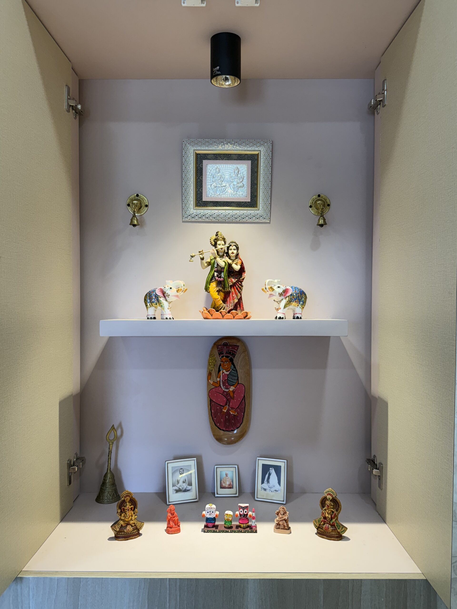 Modern built-in home mandir with a soft pink background, featuring a floating shelf with a Radha Krishna idol, decorative brass bells, focused ceiling spotlighting, and sacred figurines arranged neatly inside a cabinet structure.