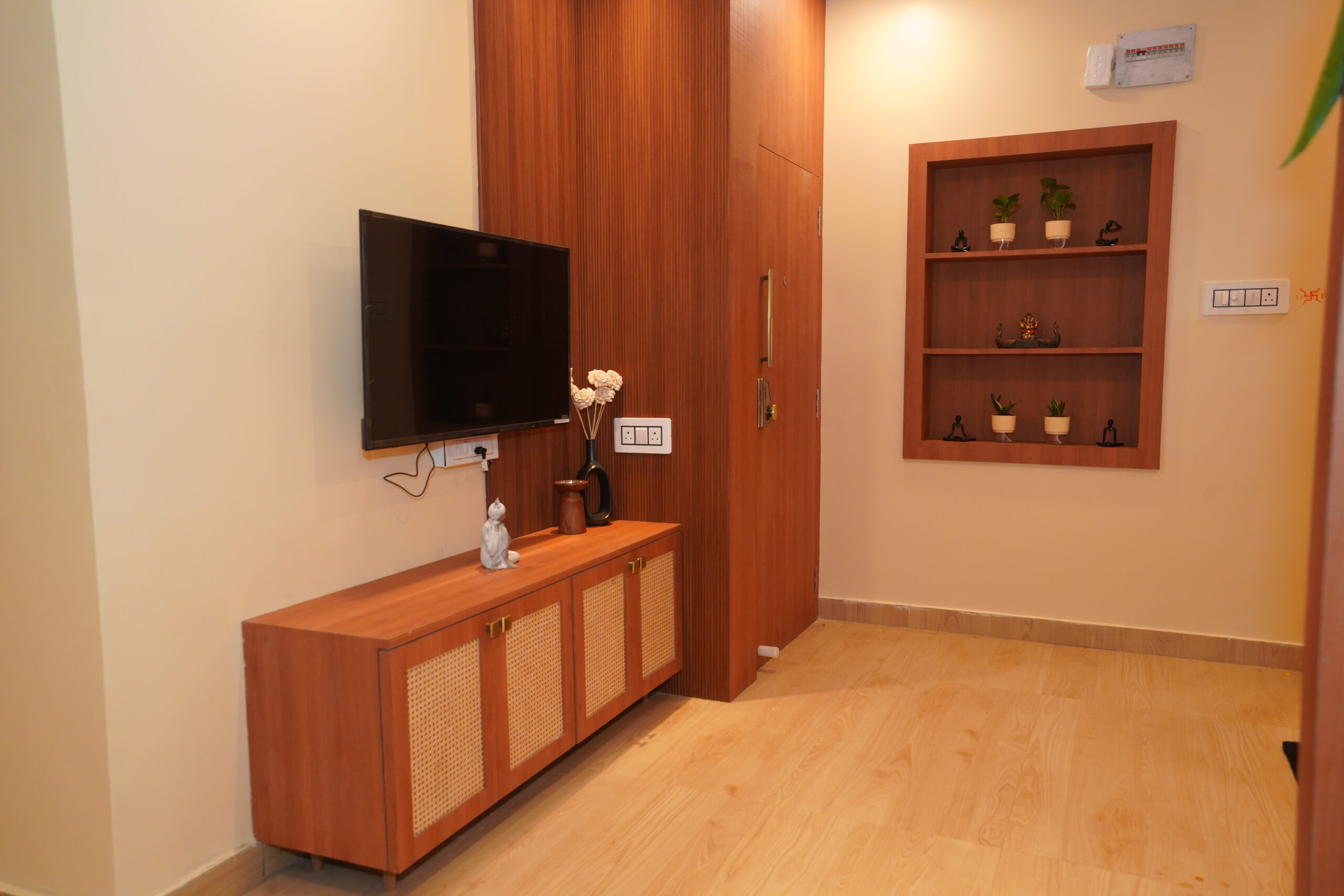 Modern living room corner featuring a wall-mounted TV over a warm wood console unit with woven cane shutter inserts, a full-height wooden wall panel camouflaging a doorway, and an adjacent built-in open display niche with small potted plants