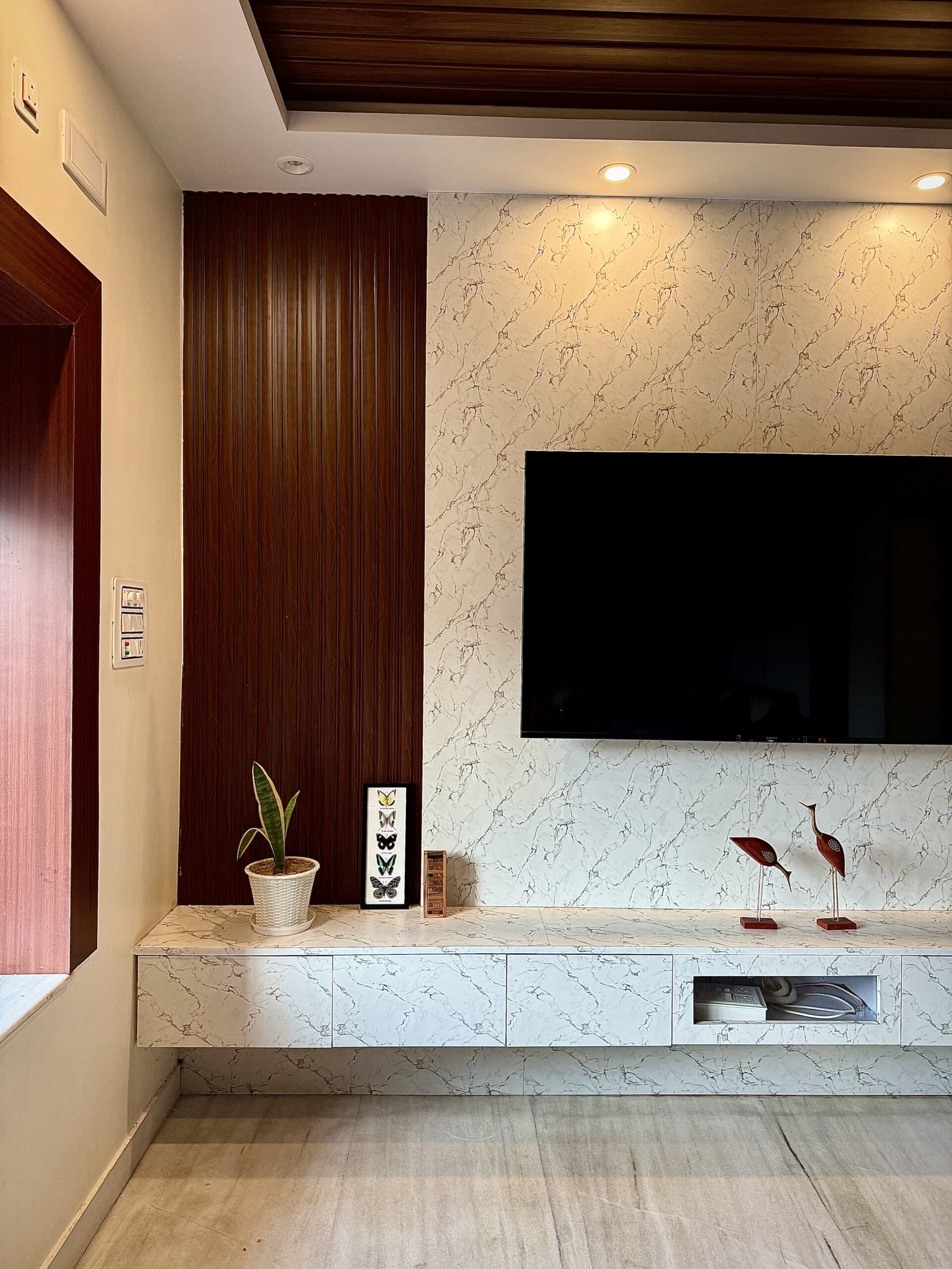 Modern entertainment center featuring a wall-mounted TV on a white marble-patterned accent panel, an asymmetrical dark wood fluted wall panel section, a matching marble-textured floating media console, and a wooden rafter false ceiling with warm spotlights