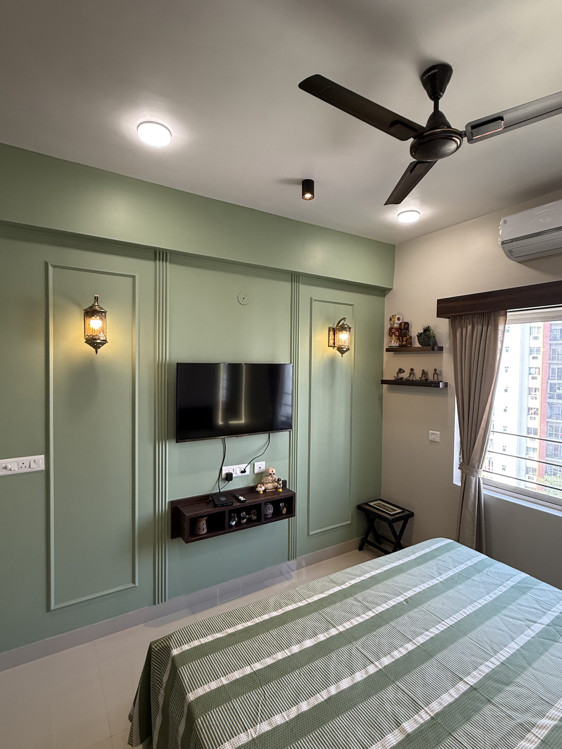 Bedroom interior featuring a sage green accent TV wall with classic wall moulding paneling, wall-mounted television, a floating wooden shelf, and traditional illuminated wall sconces.