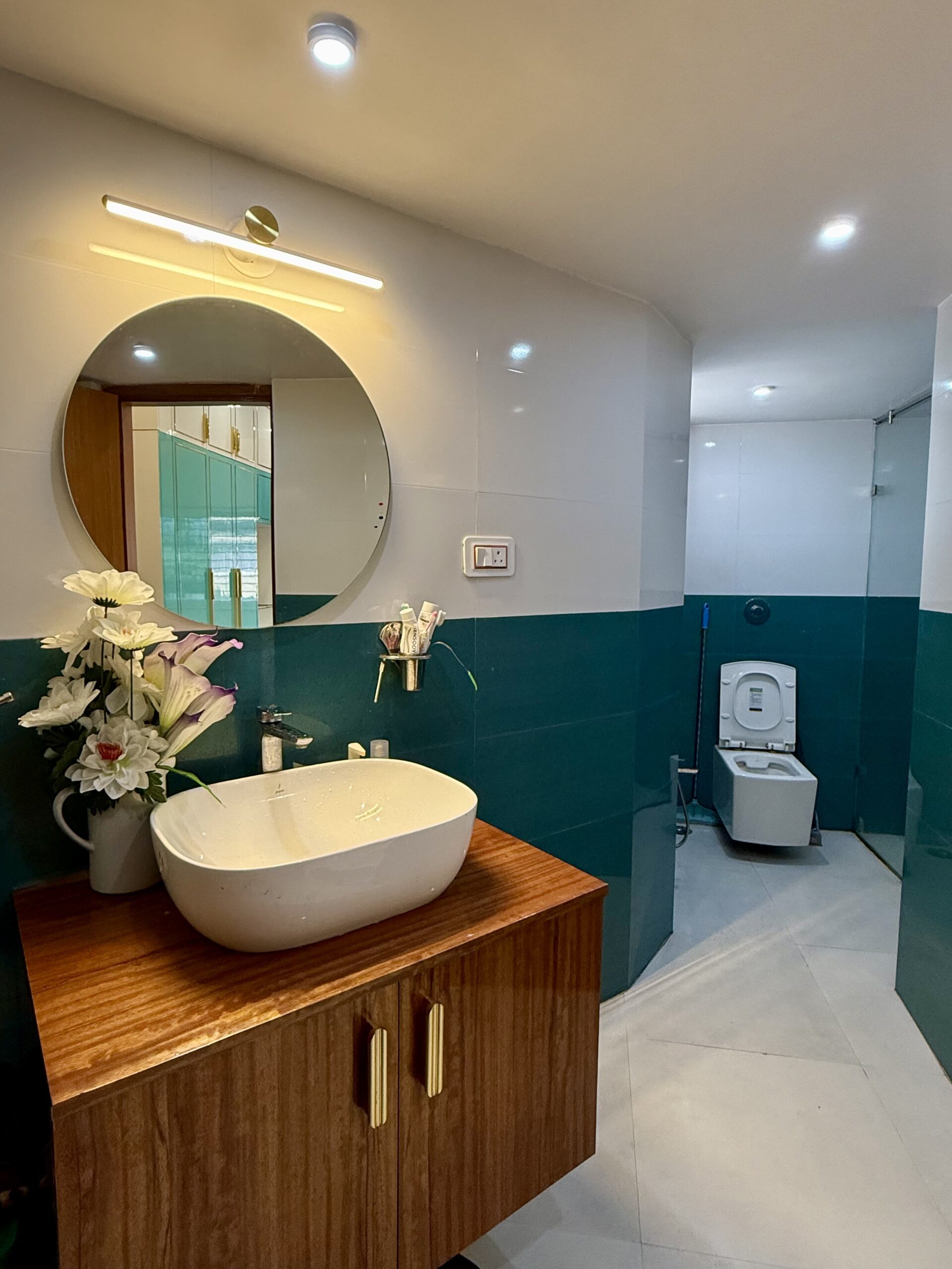 Modern bathroom interior featuring horizontal teal and white color-blocked wall tiles, a rich grain wooden vanity console with gold handles, a white tabletop washbasin, a circular wall mirror with a brass linear light fixture, and a wall-hung commode in the background