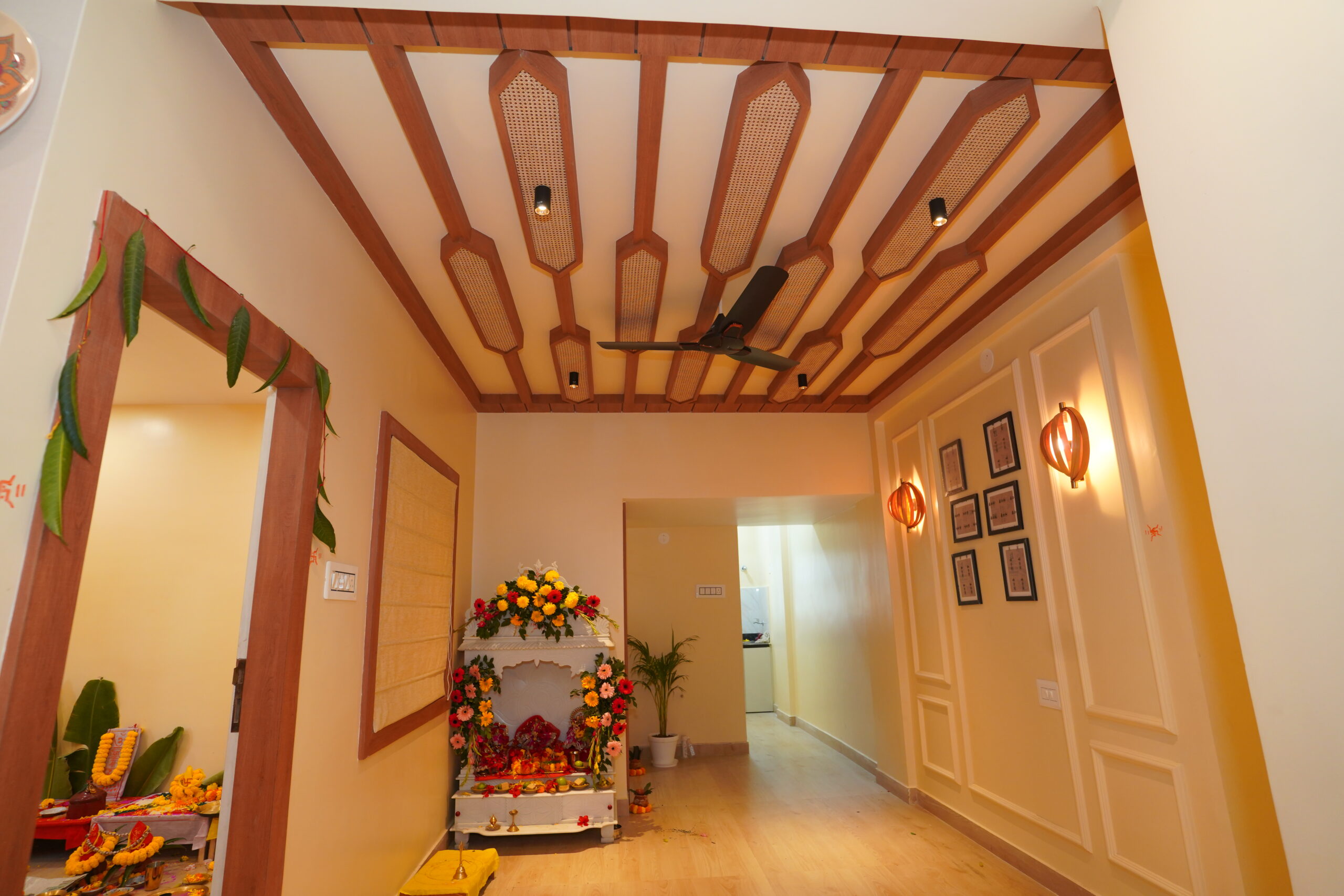 Residential hallway featuring a traditional white marble temple setup, an intricate false ceiling with wooden rafters and woven cane panel inserts, a gallery wall with classic cream moldings, and warm decorative wall sconces